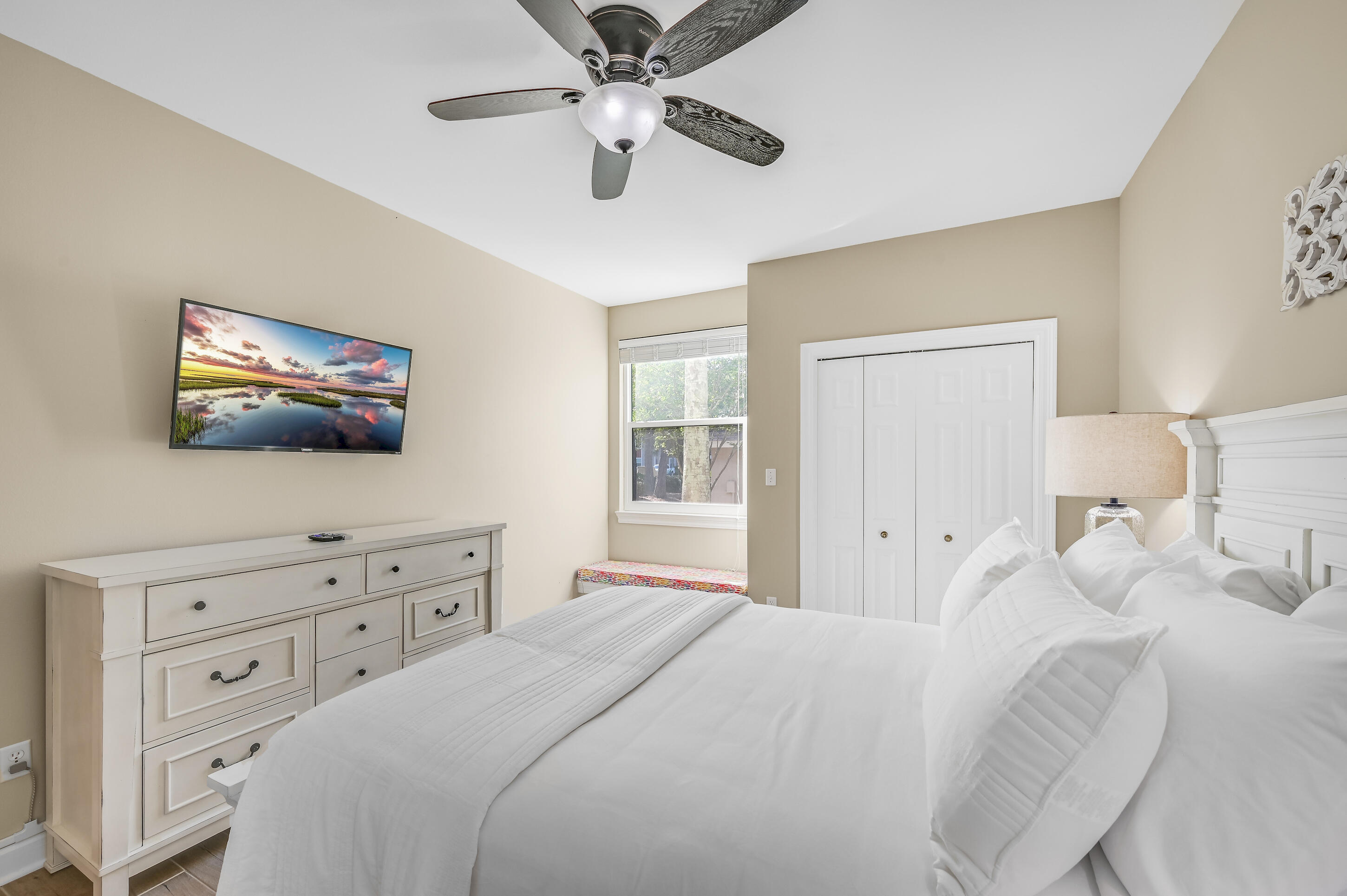 Furnished bedroom with a white bed, ceiling fan, dresser, wall-mounted TV, and window.