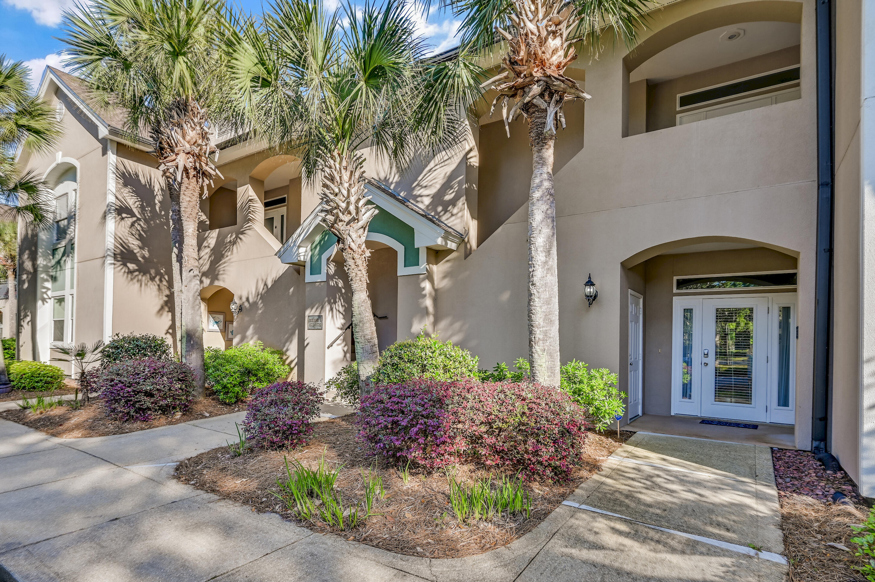 Beige residence entrance with white double doors, palm trees, and landscaped shrubs.