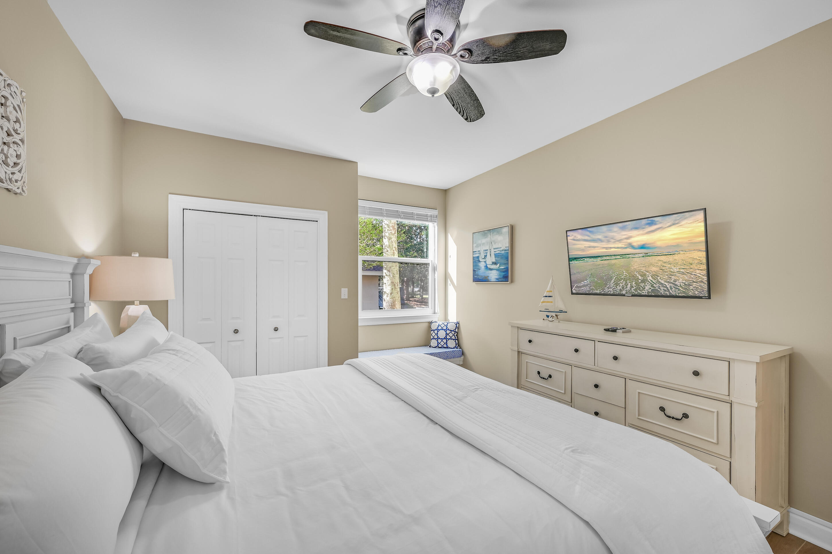 Bedroom with a white bed, ceiling fan, double-door closet, and dresser beneath coastal artwork.