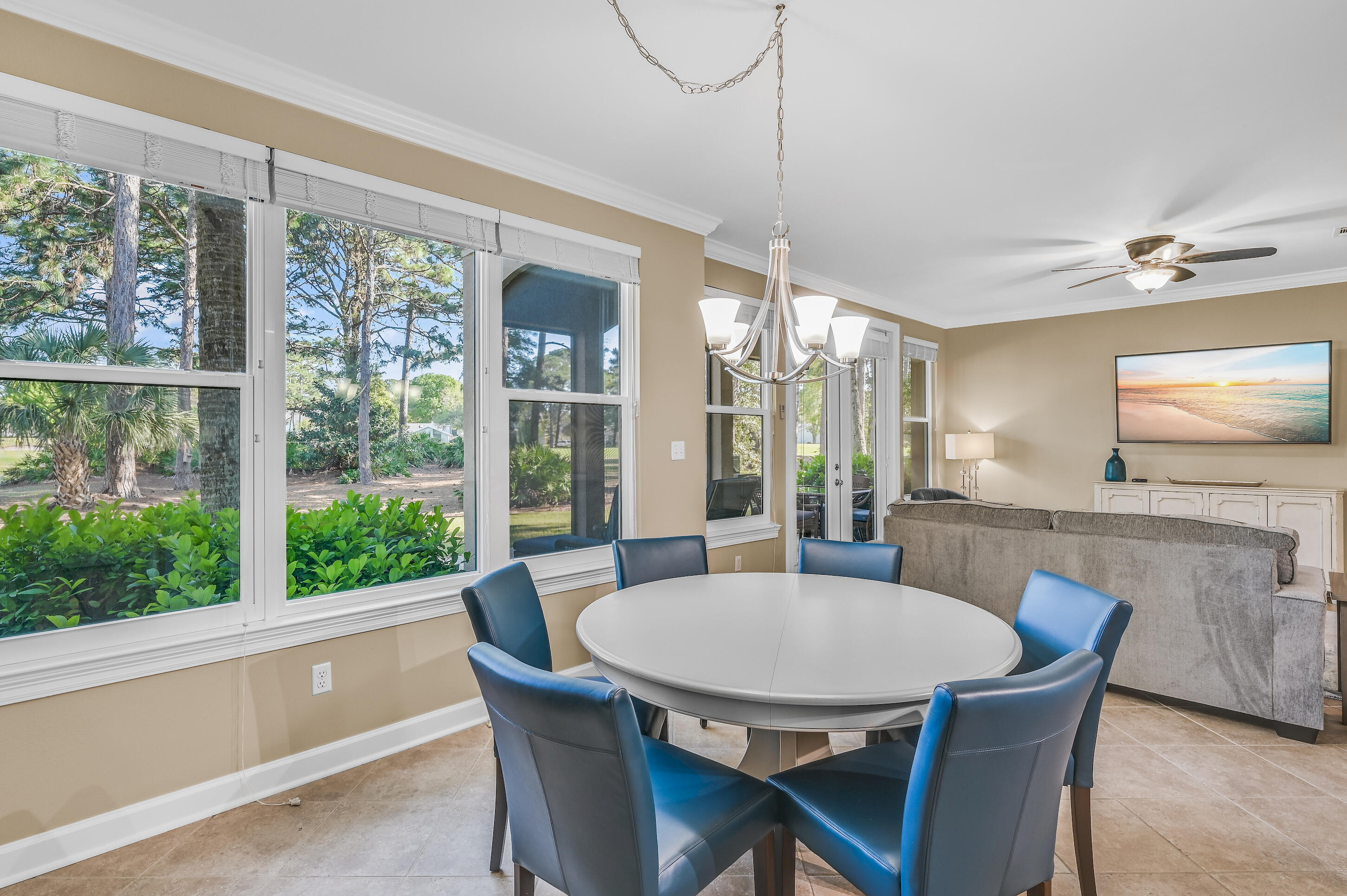 Dining and living area with a round table, blue chairs, and large windows overlooking greenery.