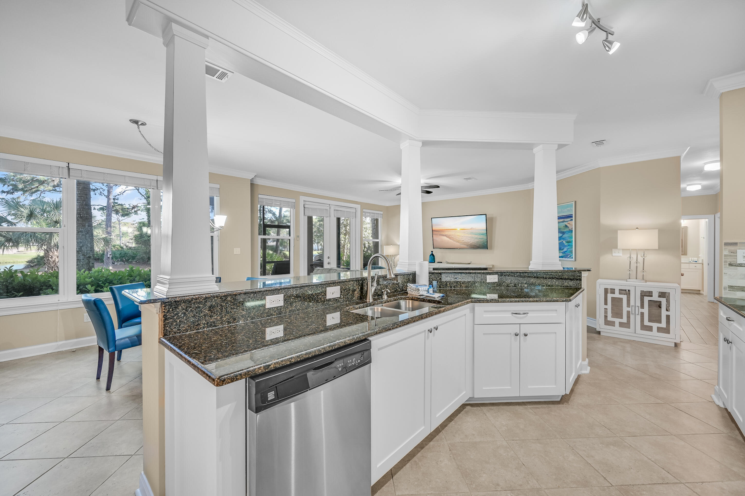 Open-plan kitchen with granite island and stainless dishwasher, beside large windows overlooking leafy landscaping.
