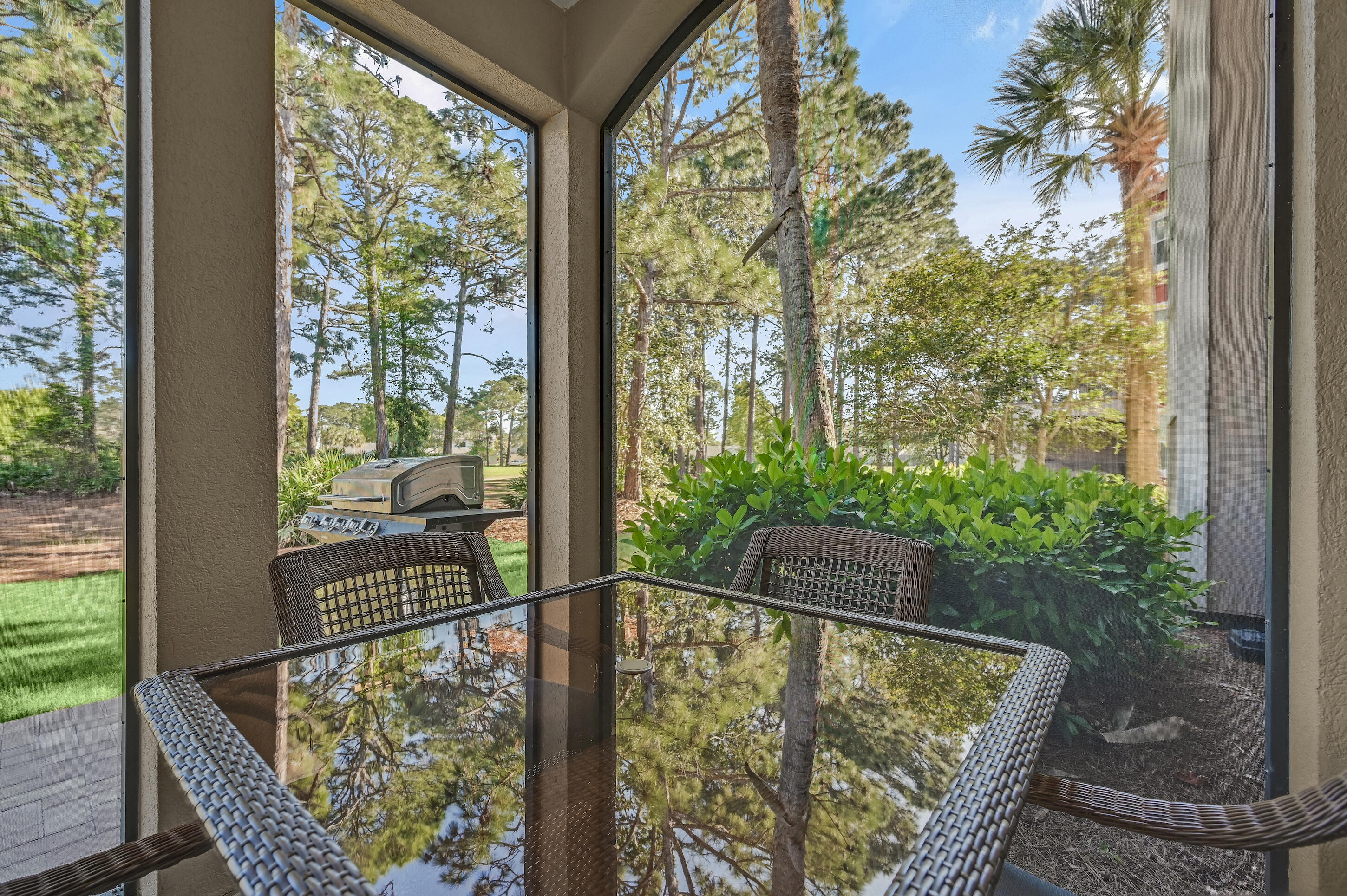 Screened patio with a wicker dining set, leafy yard, and barbecue grill.