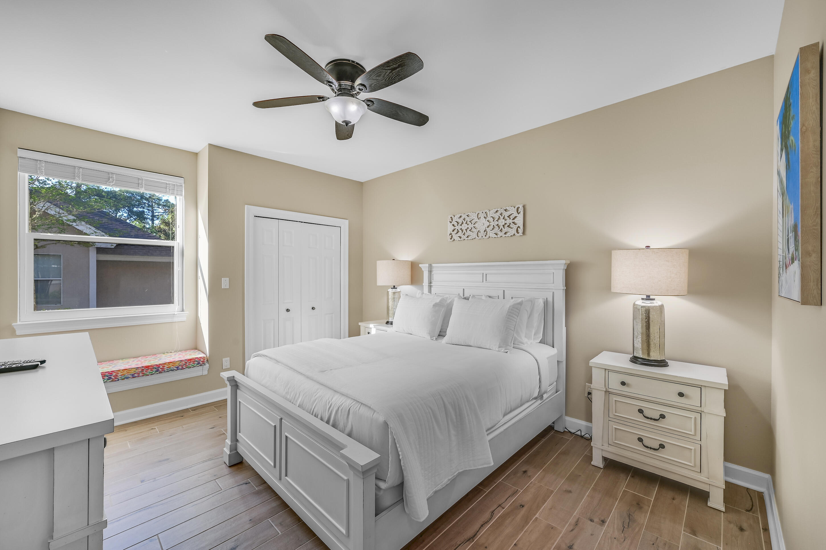 Furnished bedroom with a white paneled bed, cushioned window seat, and ceiling fan.