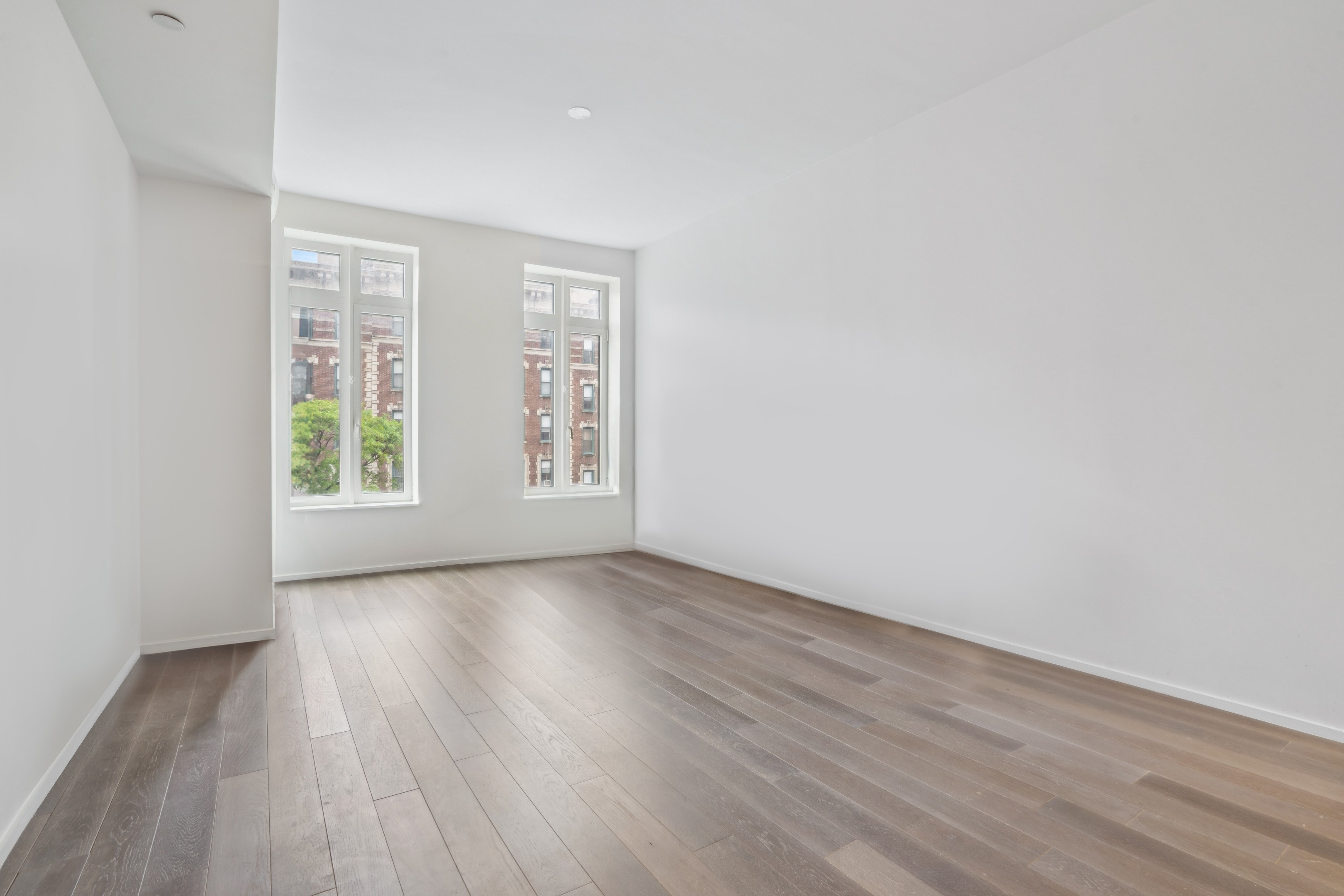 Vacant room with wide-plank wood floors and two tall windows overlooking brick buildings.
