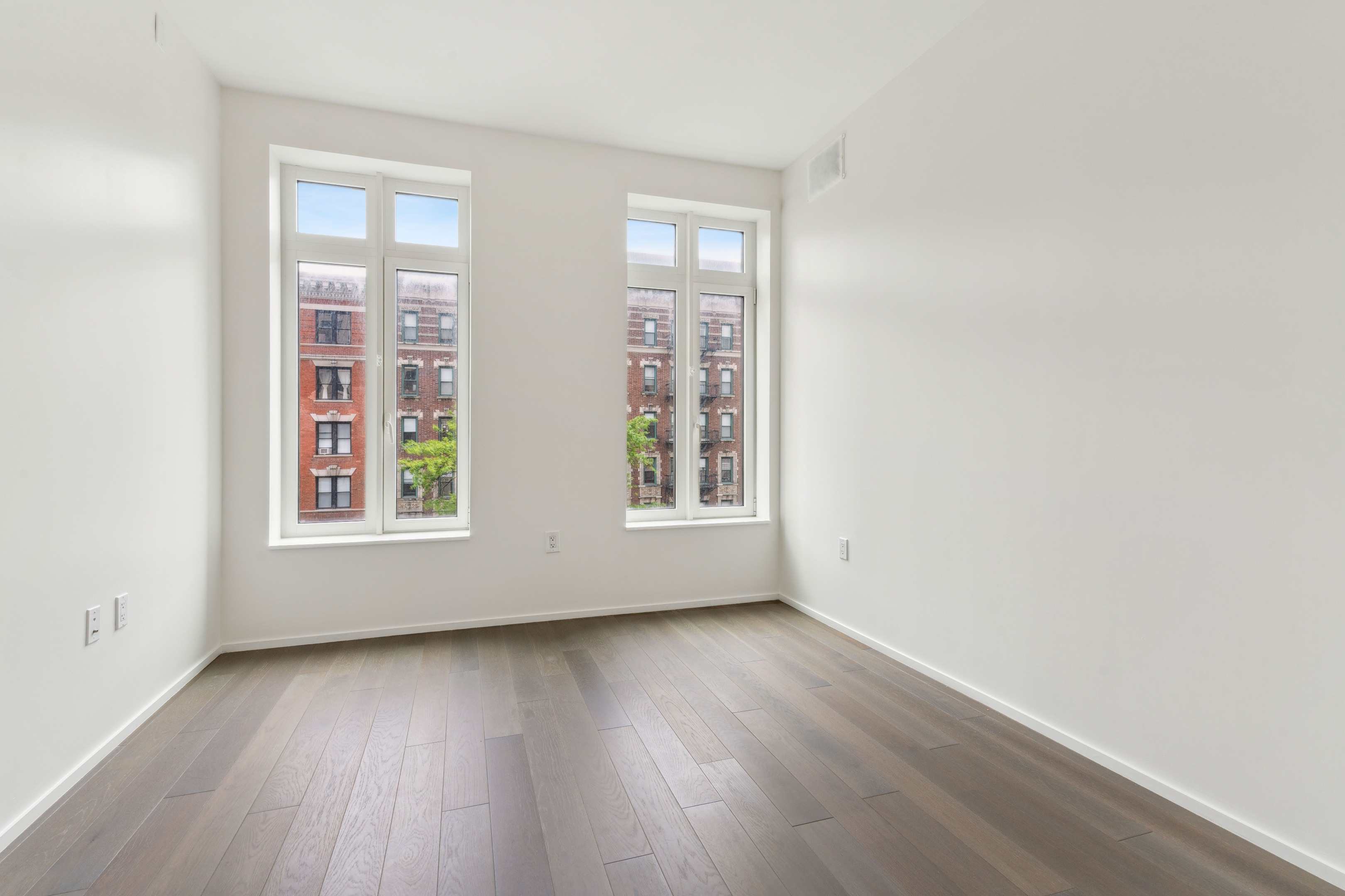 Empty room with wood plank flooring and two tall windows overlooking brick buildings.