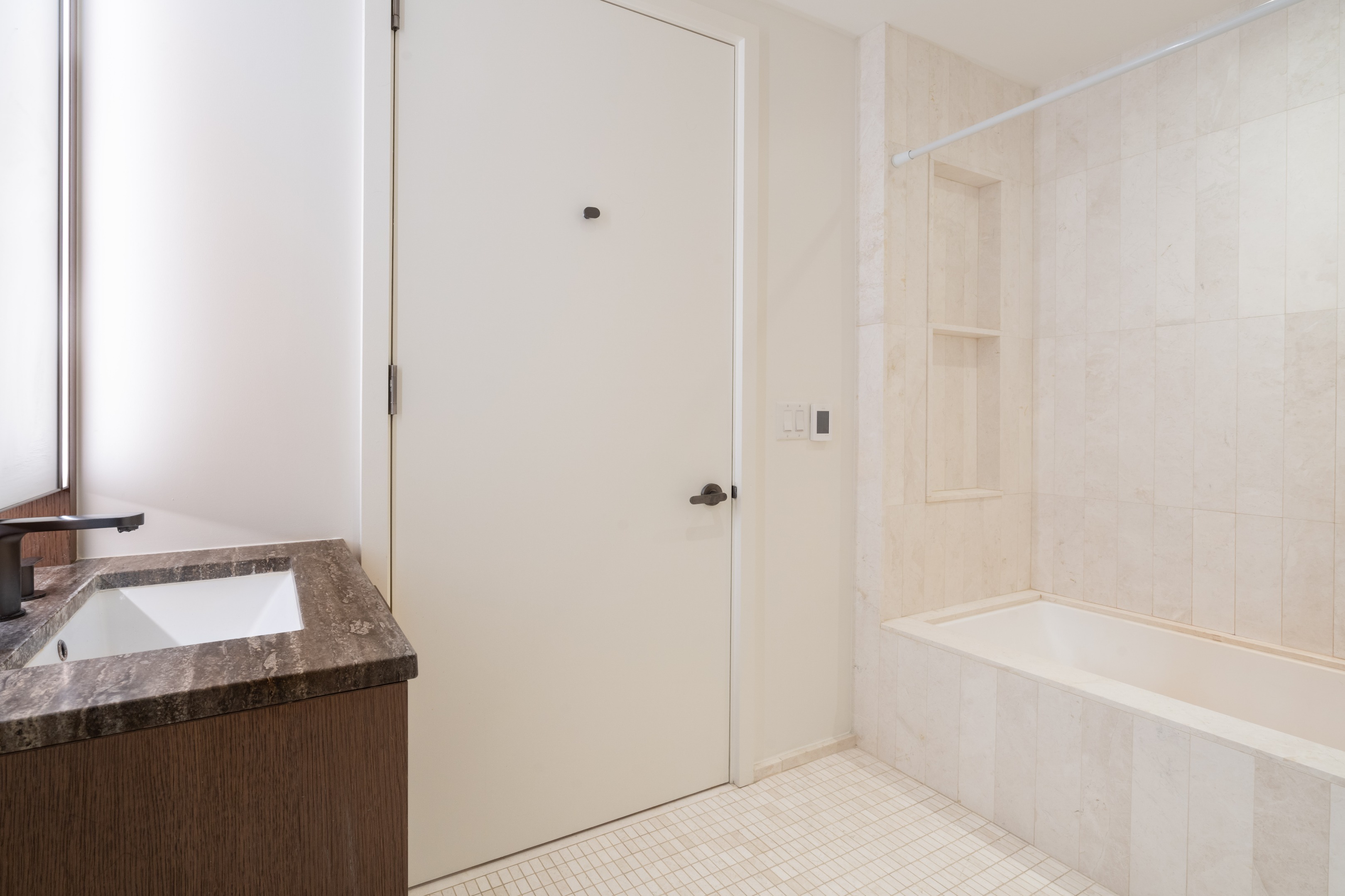 Bathroom with a pale stone-tiled bathtub and recessed shelves beside a dark stone vanity.