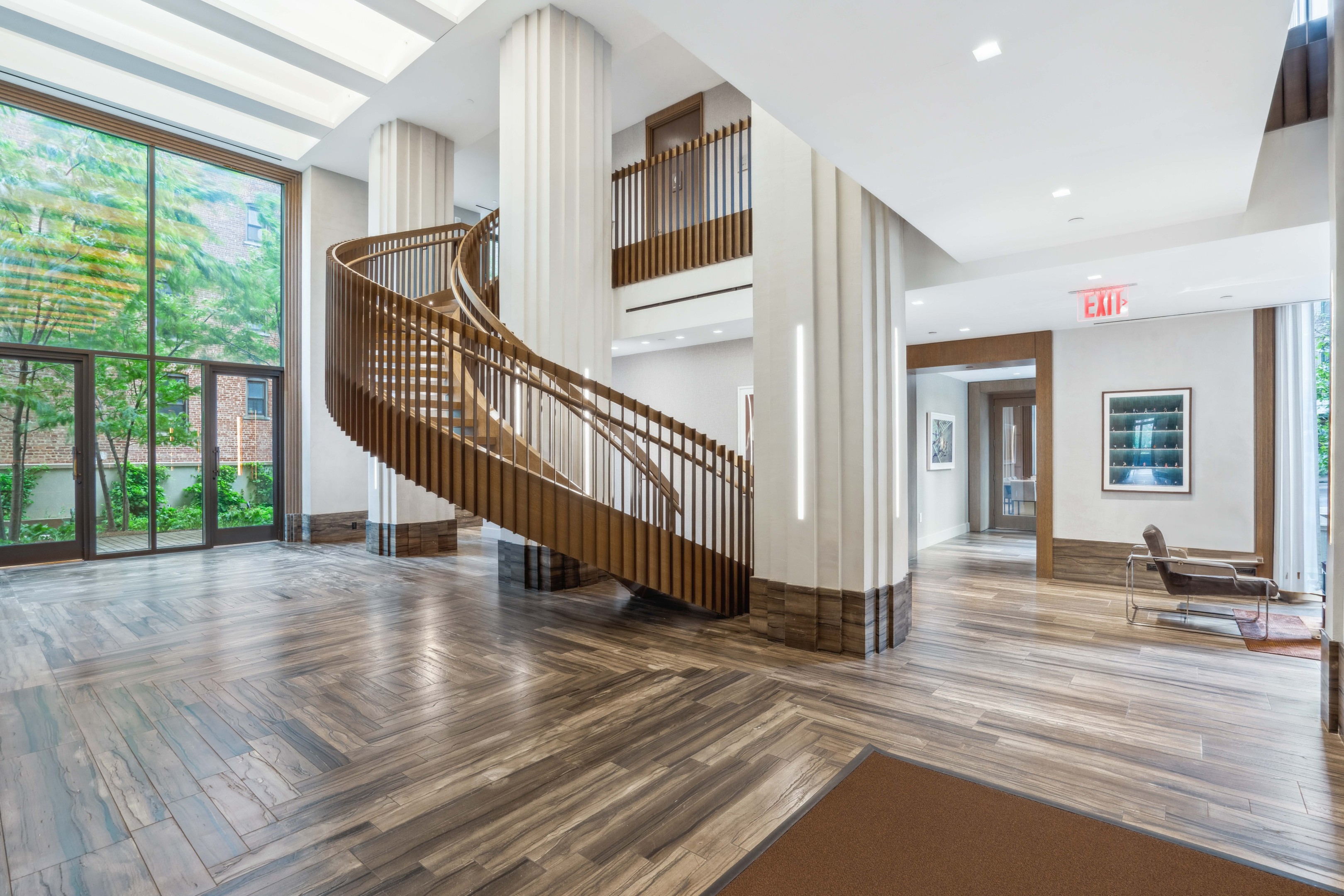 Residential lobby with a sweeping wooden staircase, tall windows, and patterned stone flooring.