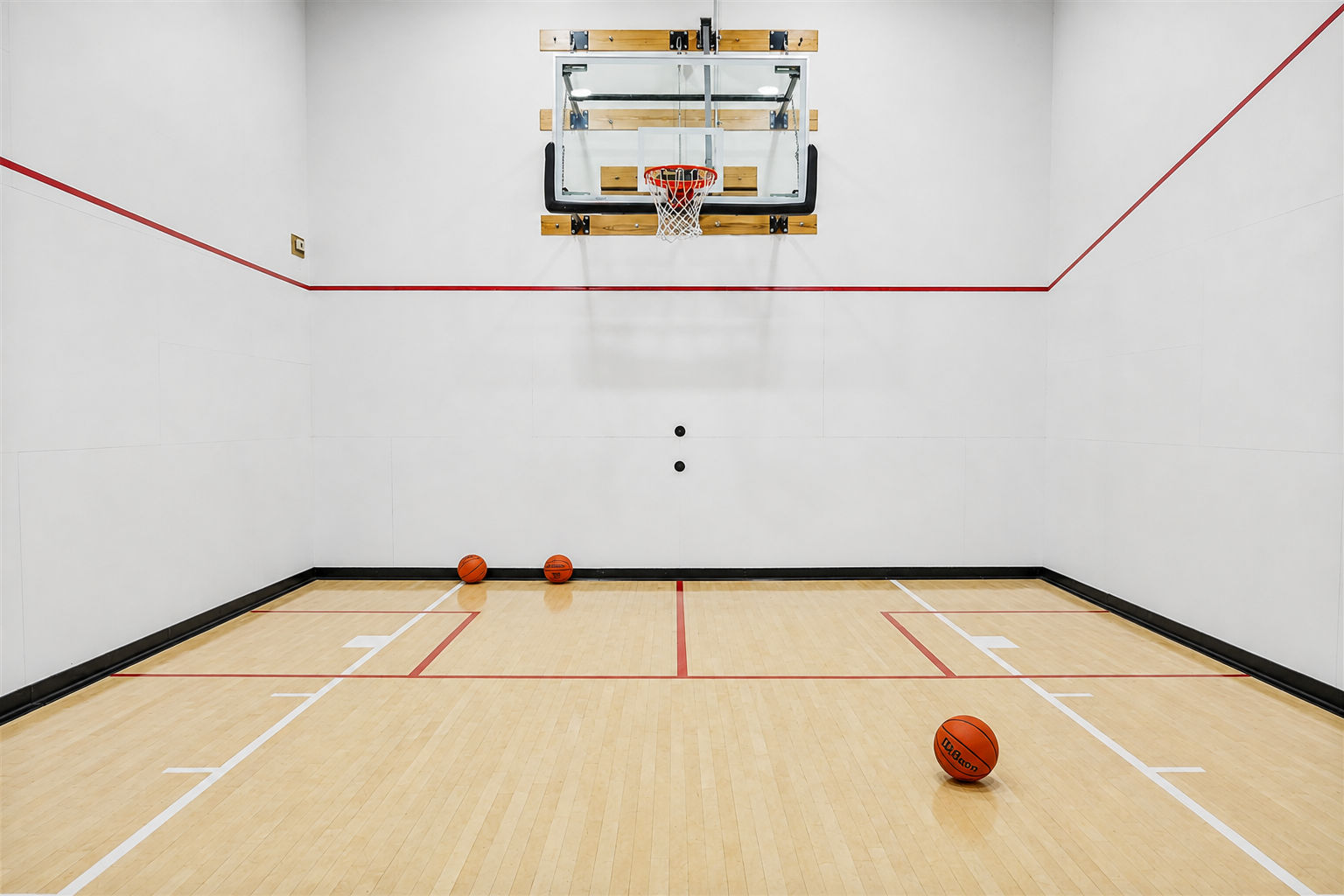 Indoor basketball court with a wall-mounted hoop and three basketballs on the hardwood floor.
