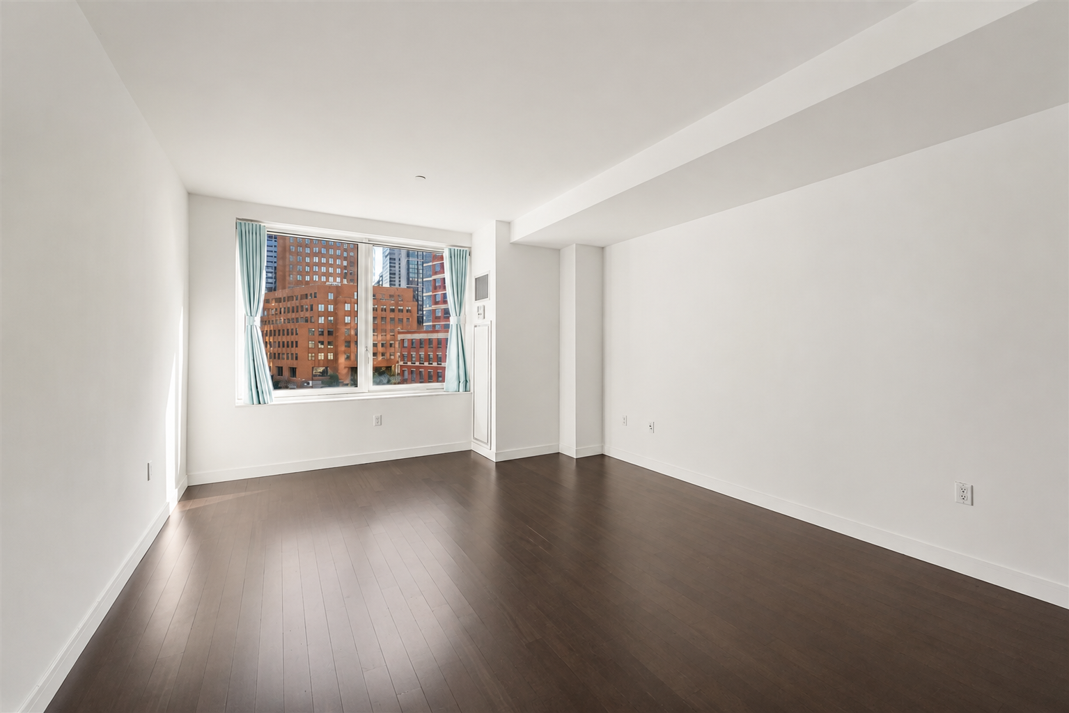 Empty apartment room with dark hardwood floors and a large curtained window overlooking city buildings.