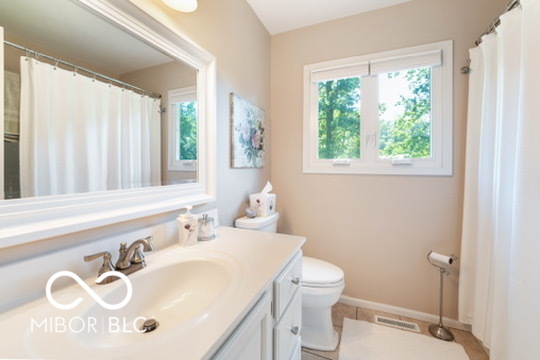 Bathroom with a white vanity and large mirror, toilet, and window overlooking leafy trees.
