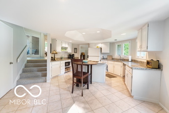 Kitchen with a central island and two stools, white cabinetry, tile flooring, and an adjoining stairway.