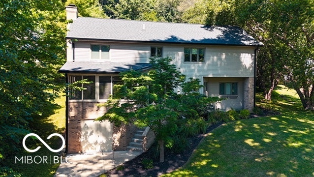 Two-story house with light siding, a brick lower level, and mature trees surrounding the landscaped yard.