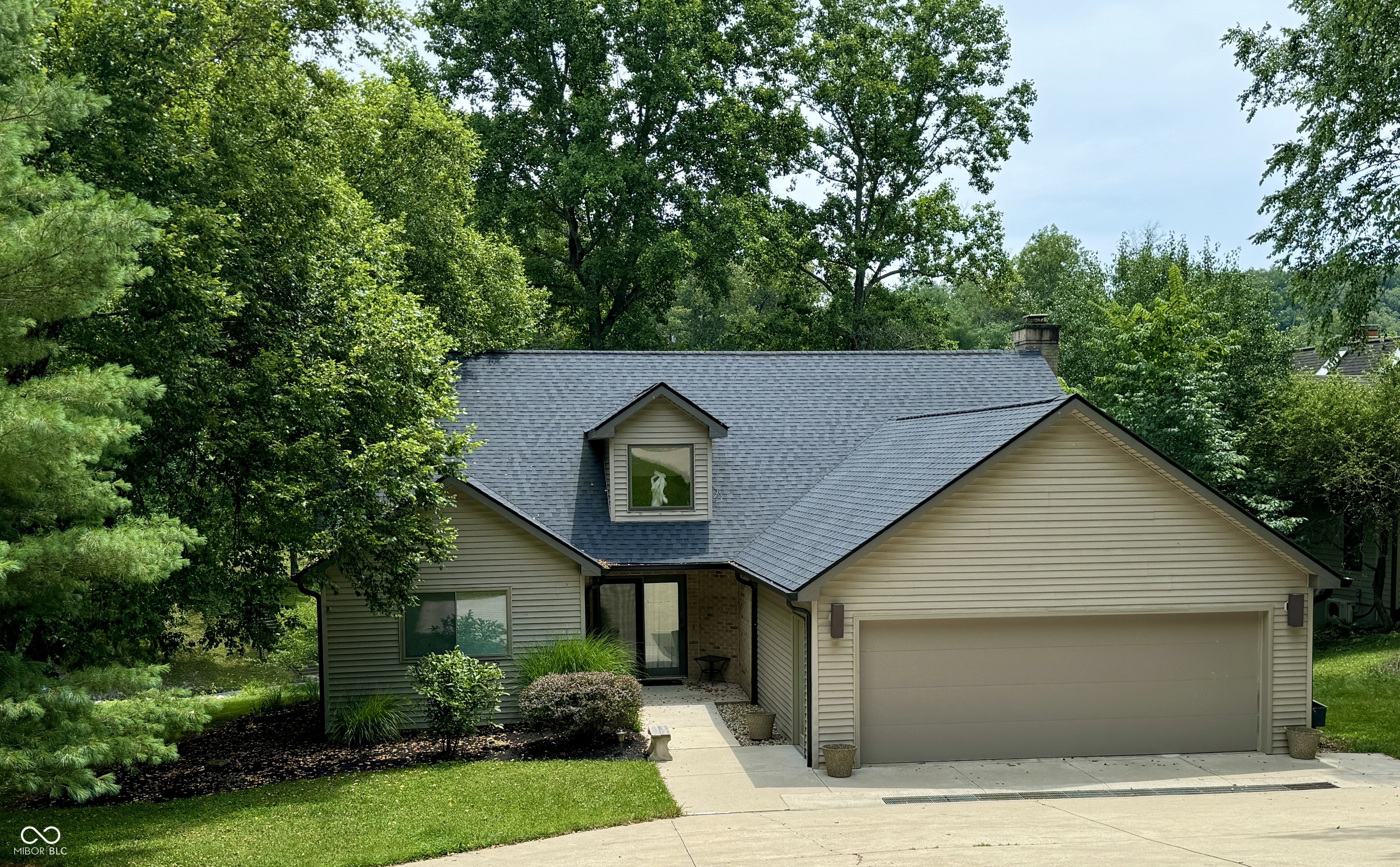 Beige home with an attached two-car garage, dark shingle roof, and mature trees.