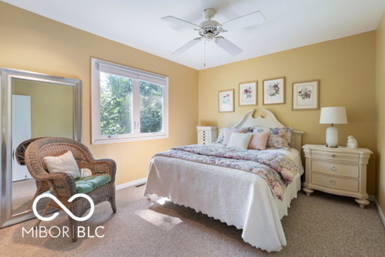 Bedroom with a white bed and floral quilt, botanical prints, and a wicker chair beside a large window.