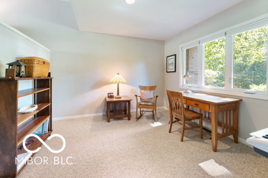 Carpeted room with a wooden desk and chairs beside wide windows overlooking trees.