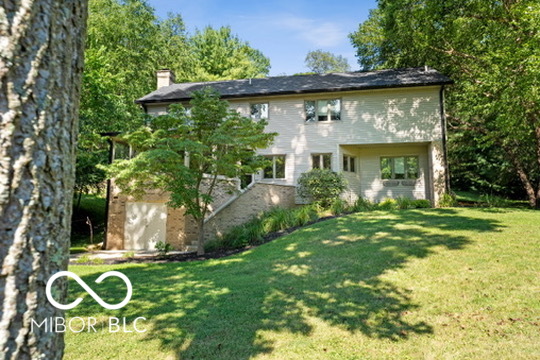 Two-story light-colored house among mature trees, with a sloping lawn.