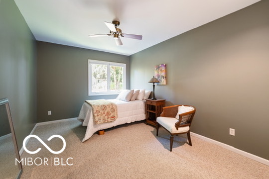 Furnished bedroom with a white bed, floral throw, and cushioned chair beside a large window.