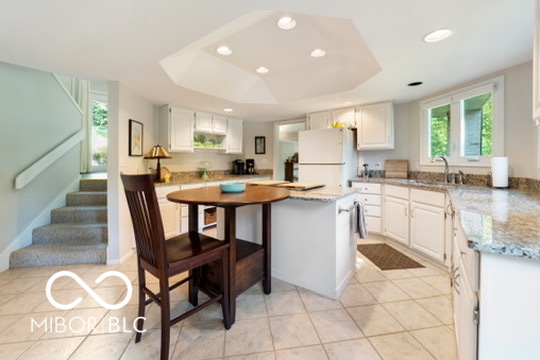 Kitchen with white cabinets and granite counters, central island and dining table, beside a carpeted staircase.