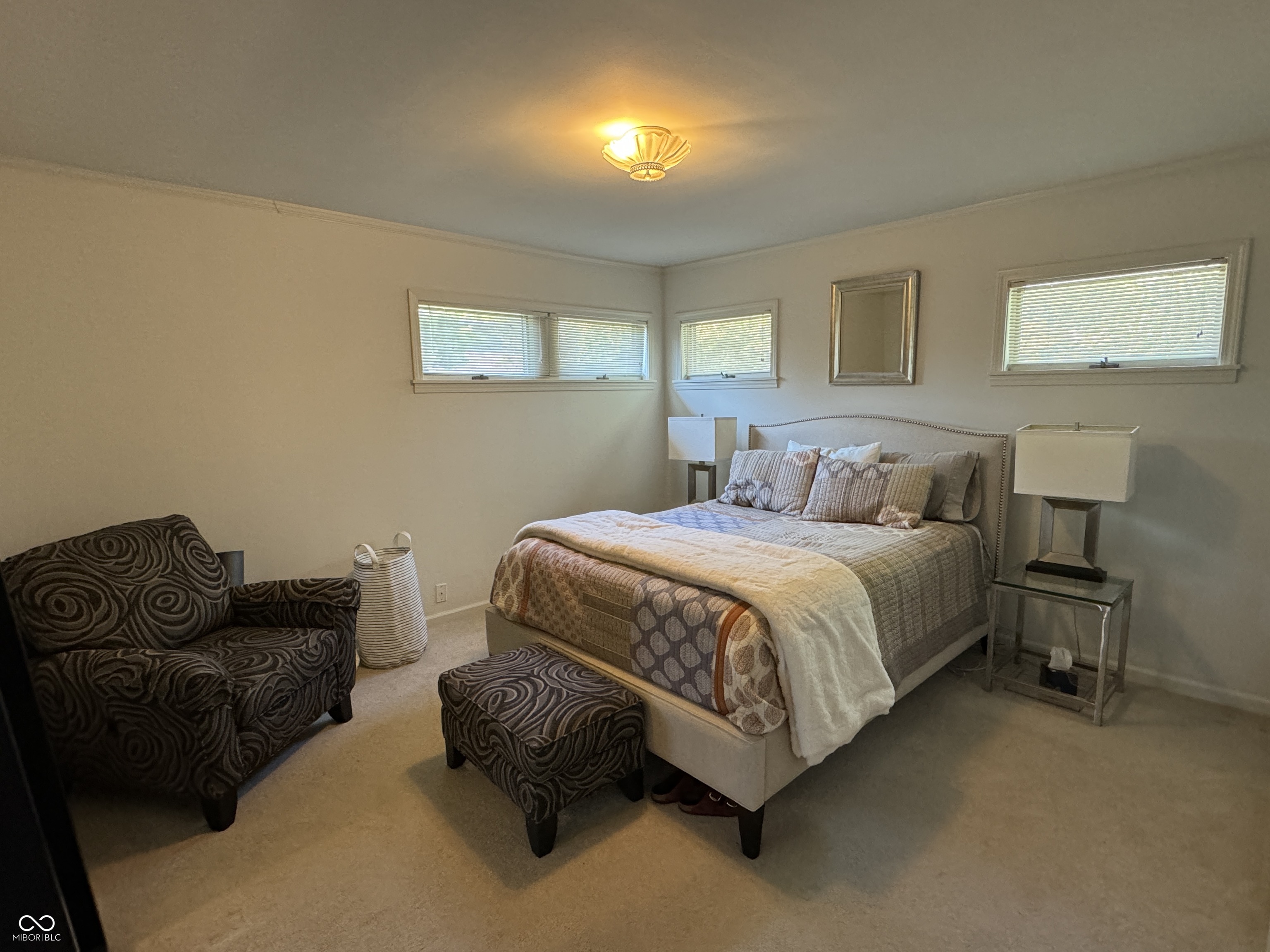 Furnished bedroom with a large bed, patterned armchair, and several high windows with blinds.