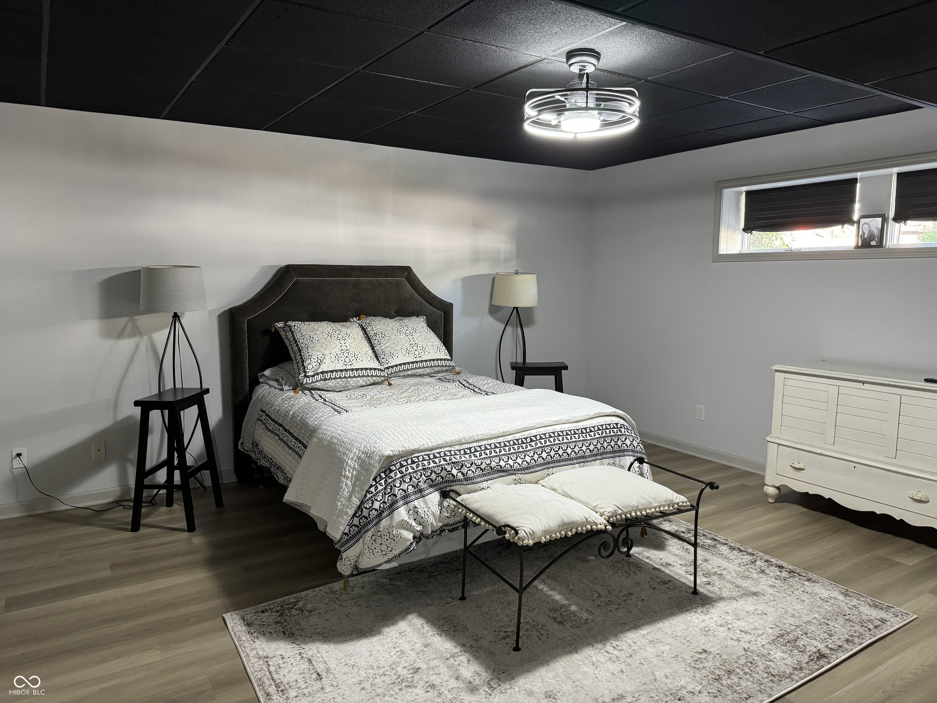 Furnished bedroom with a patterned bed, dark headboard, white dresser, and cushioned bench.