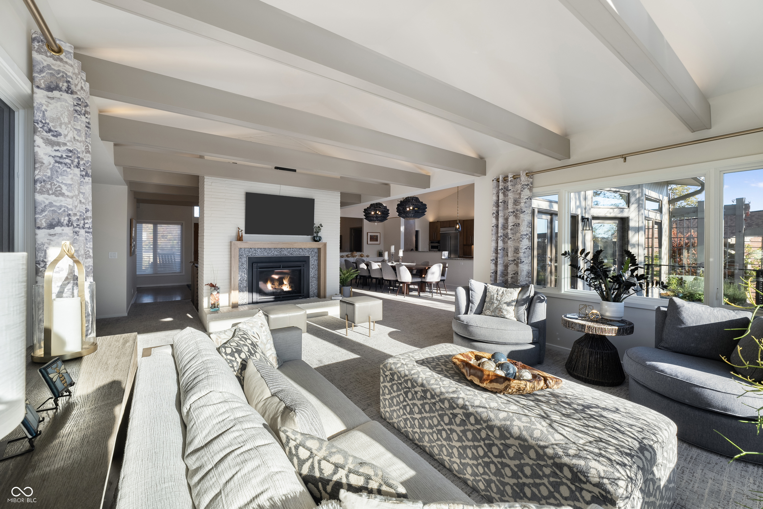 Staged living room with vaulted beamed ceiling, white brick fireplace, and large windows.