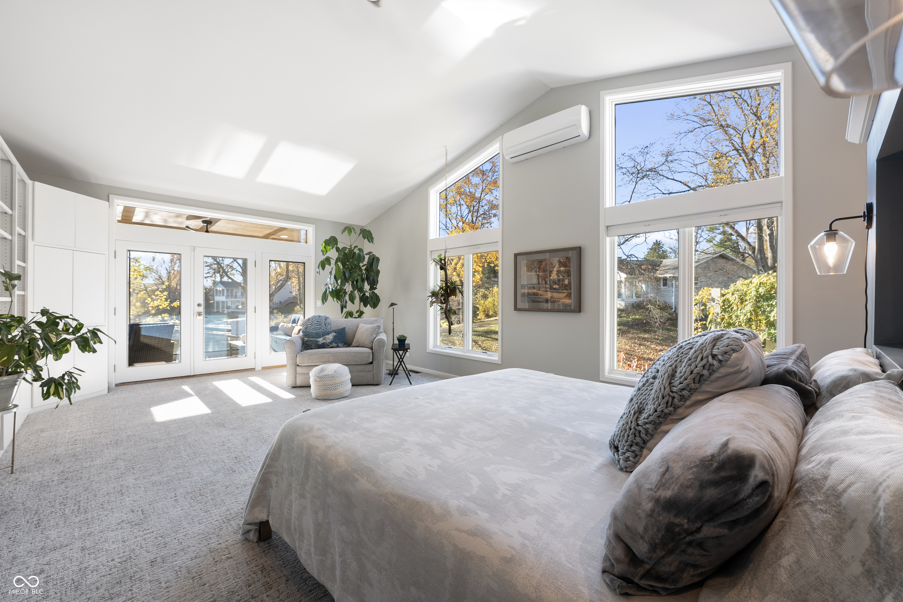 Bright bedroom with a large bed, sitting area, and tall windows overlooking autumn trees.