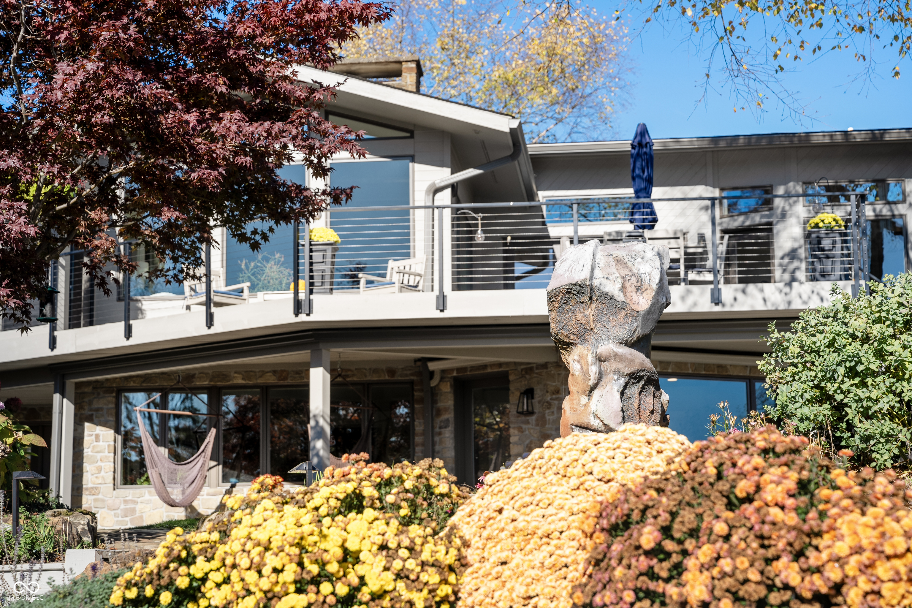 Modern home with a broad deck, garden hammock, stone sculpture, and colorful autumn flowers.