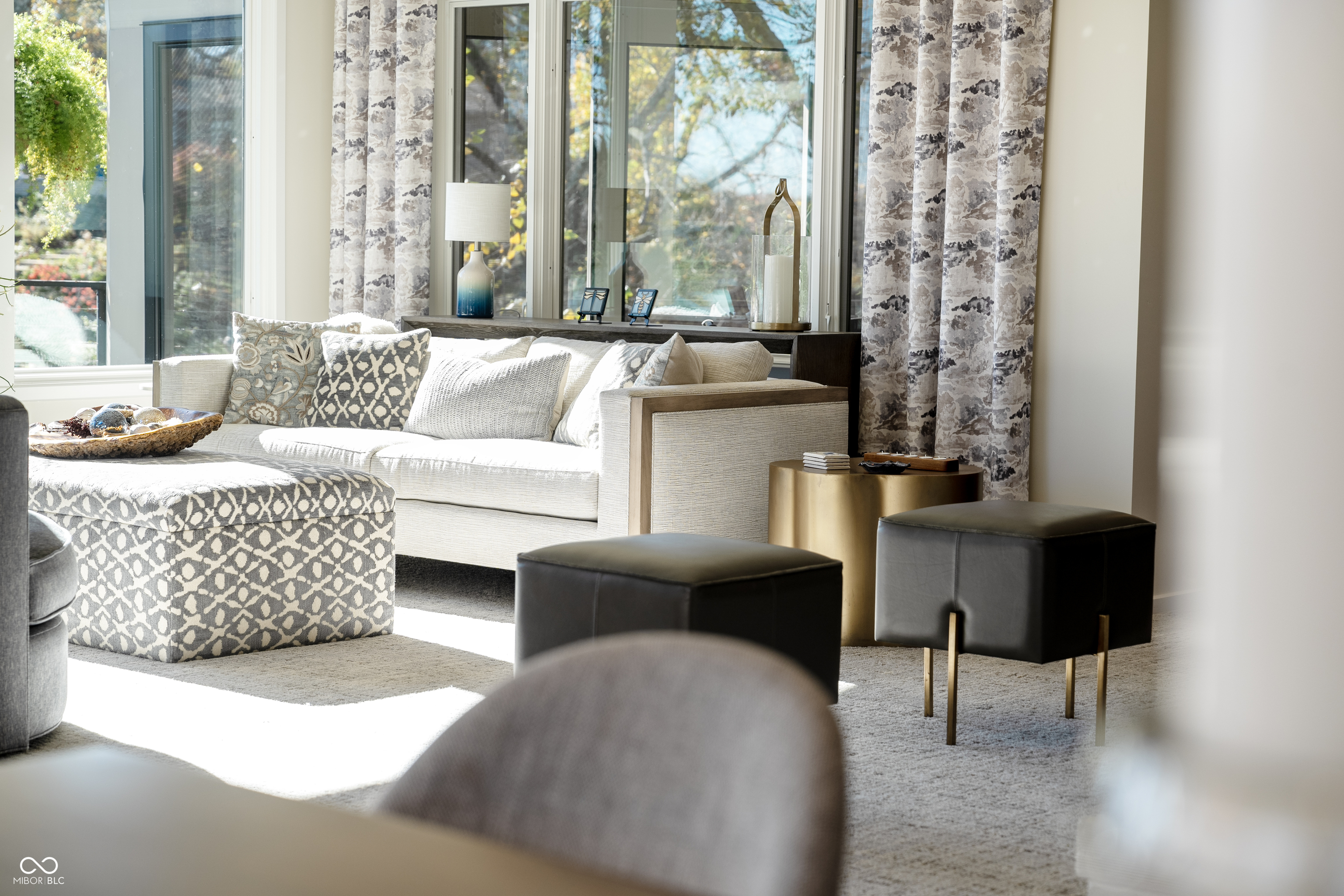 Living room with a cream sofa, patterned ottoman, dark stools, and large windows overlooking autumn foliage.