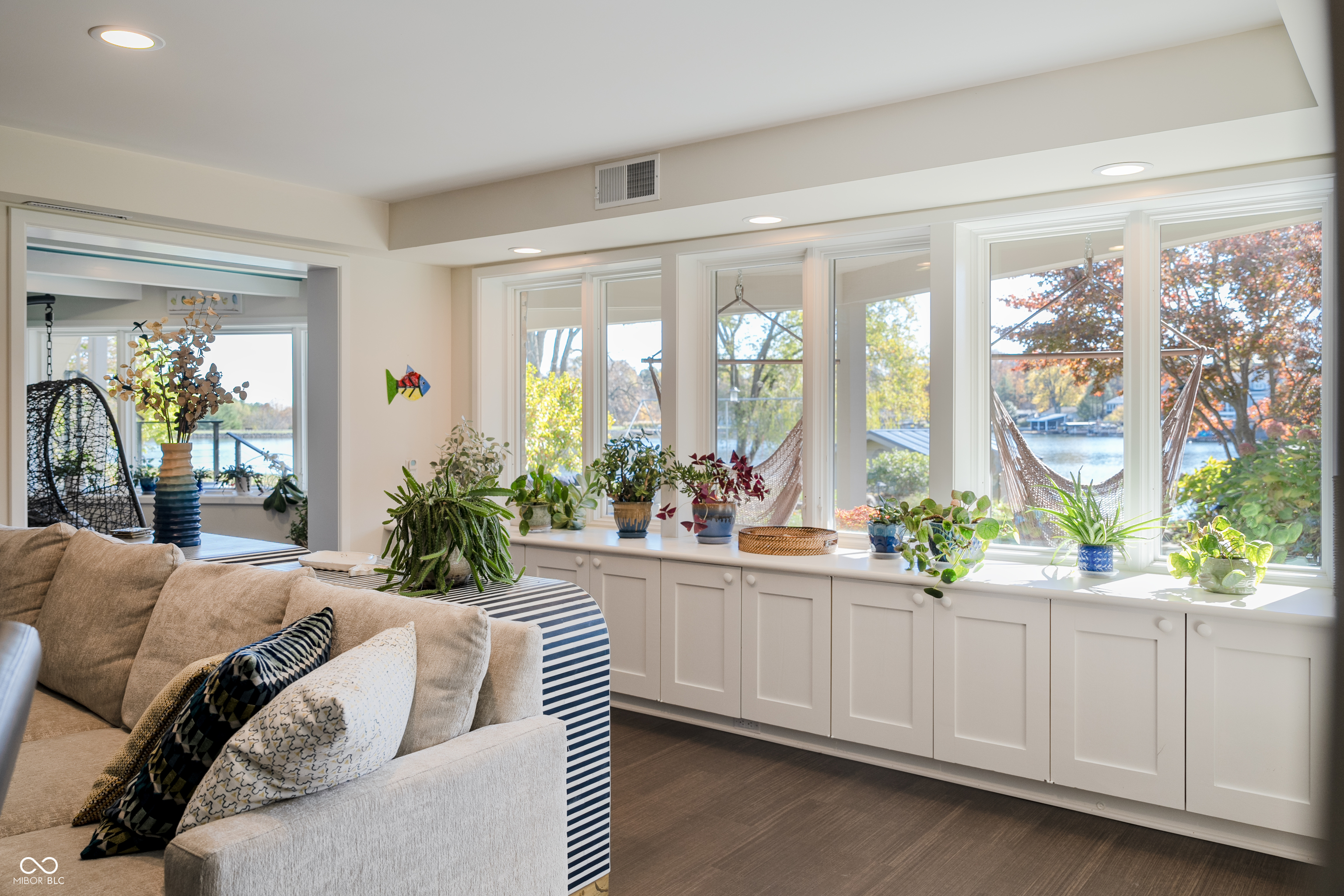Living room with lake-view windows, white built-in cabinets, indoor plants, and a beige sofa.
