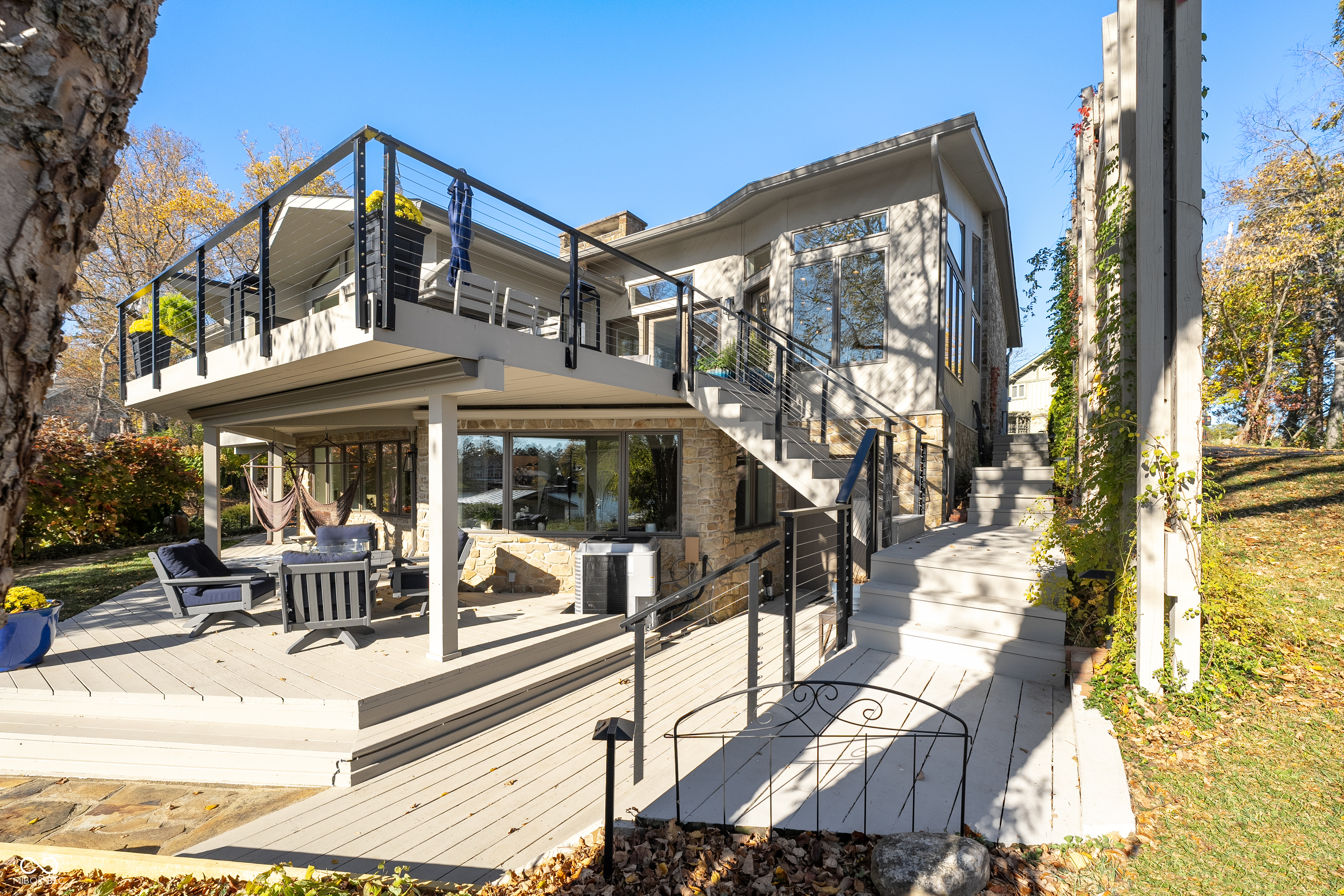 Modern house with stone lower level, multi-level decks, black railings, and exterior stairs among autumn trees.