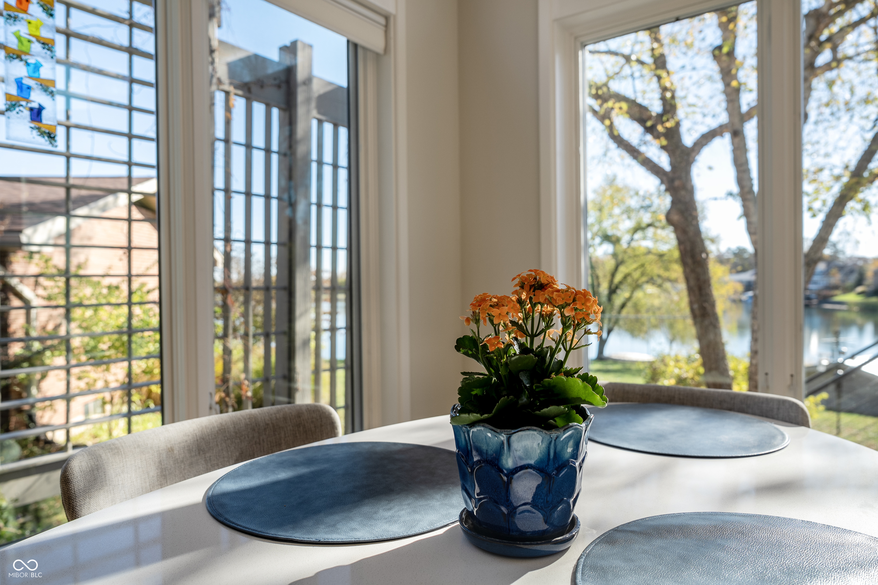 Dining table with blue placemats and orange flowers, beside windows overlooking a lake.