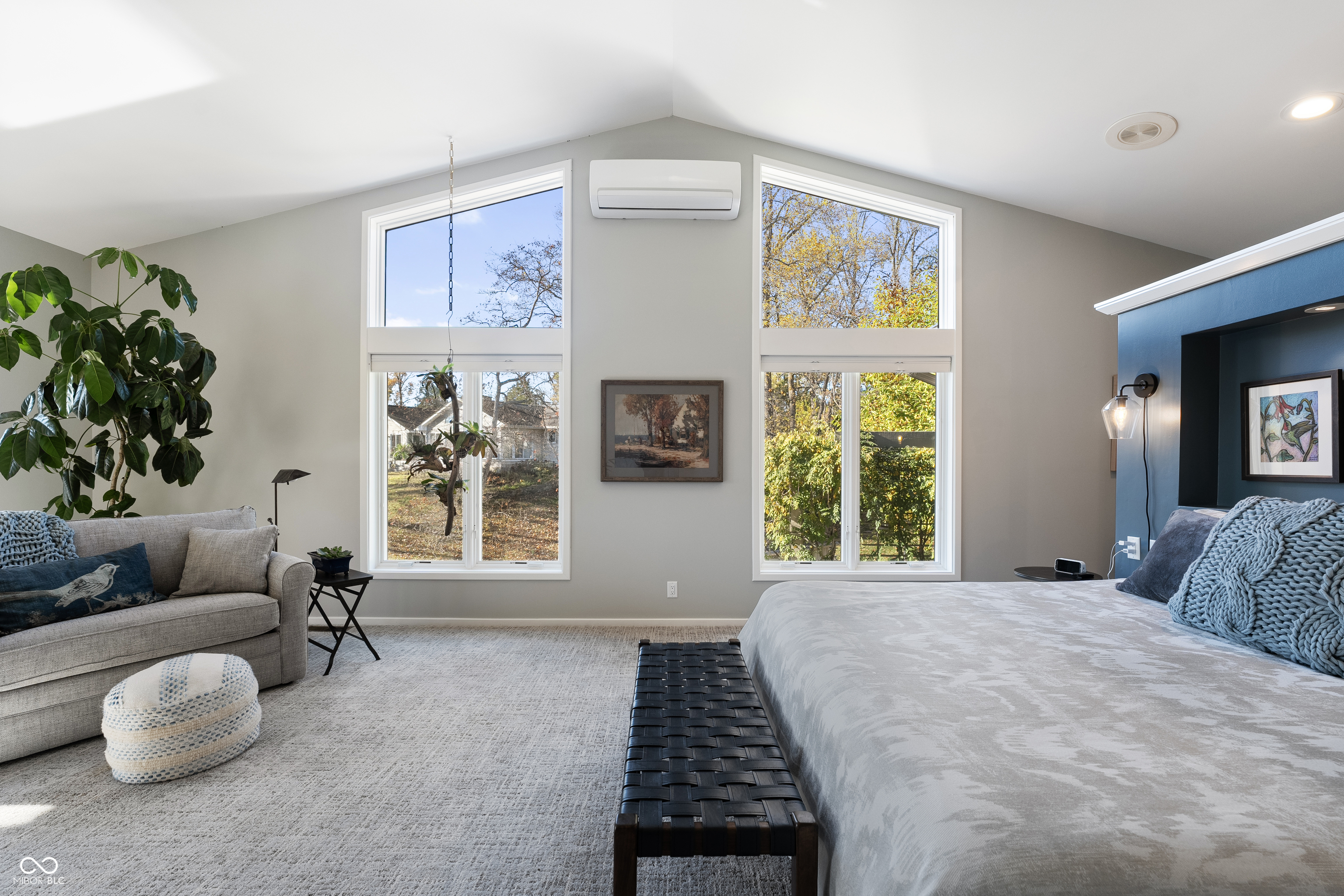 Furnished bedroom with a vaulted ceiling, tall windows, gray bed, and sofa beside a large potted plant.