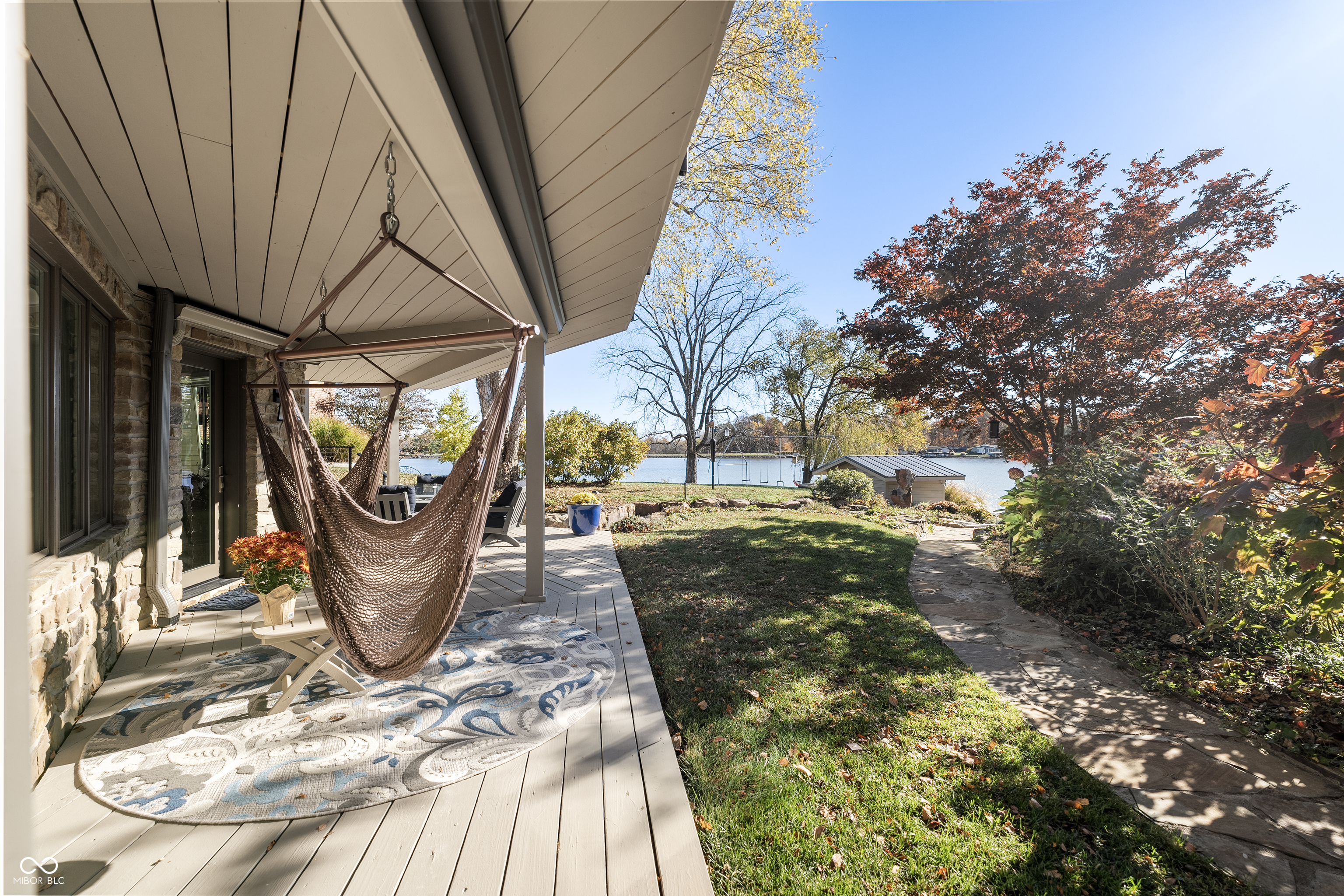 Covered lakeside patio with a hanging chair, stone path, autumn trees, and water beyond the lawn.