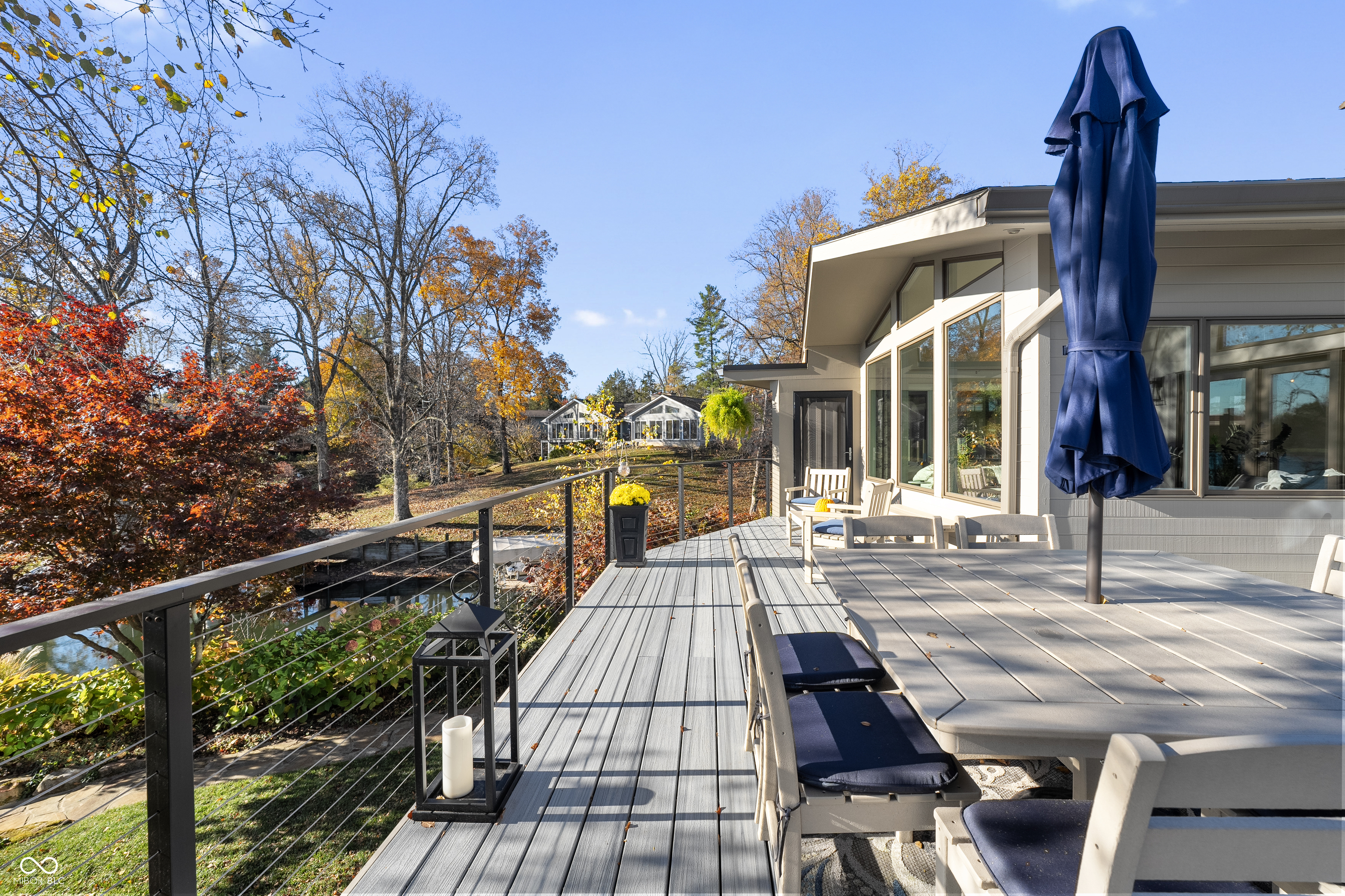 Furnished home deck with a dining table, closed navy umbrella, and autumn trees beside the water.