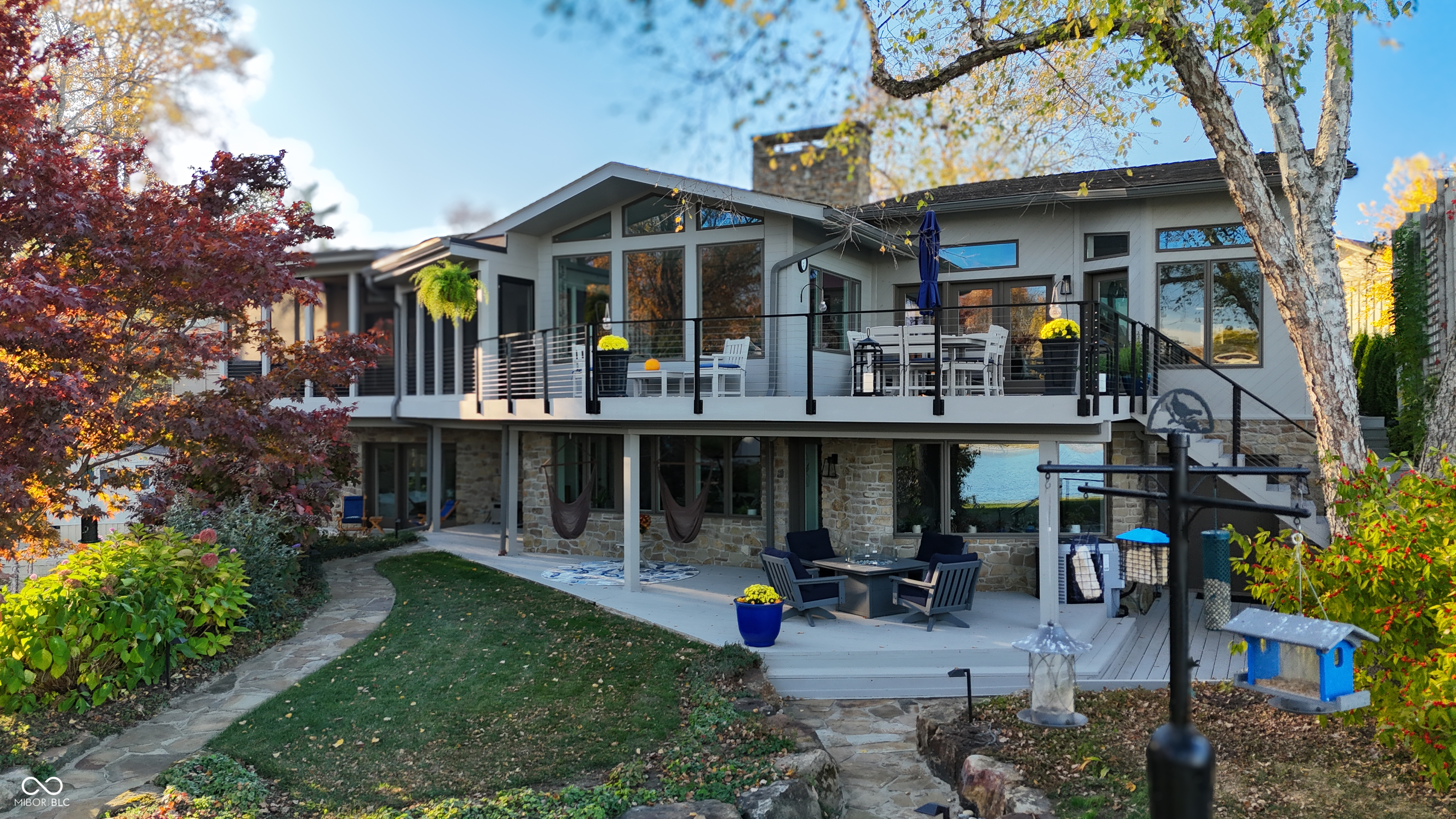 Modern lakefront home with a furnished raised deck, stone lower level, and autumn landscaping.