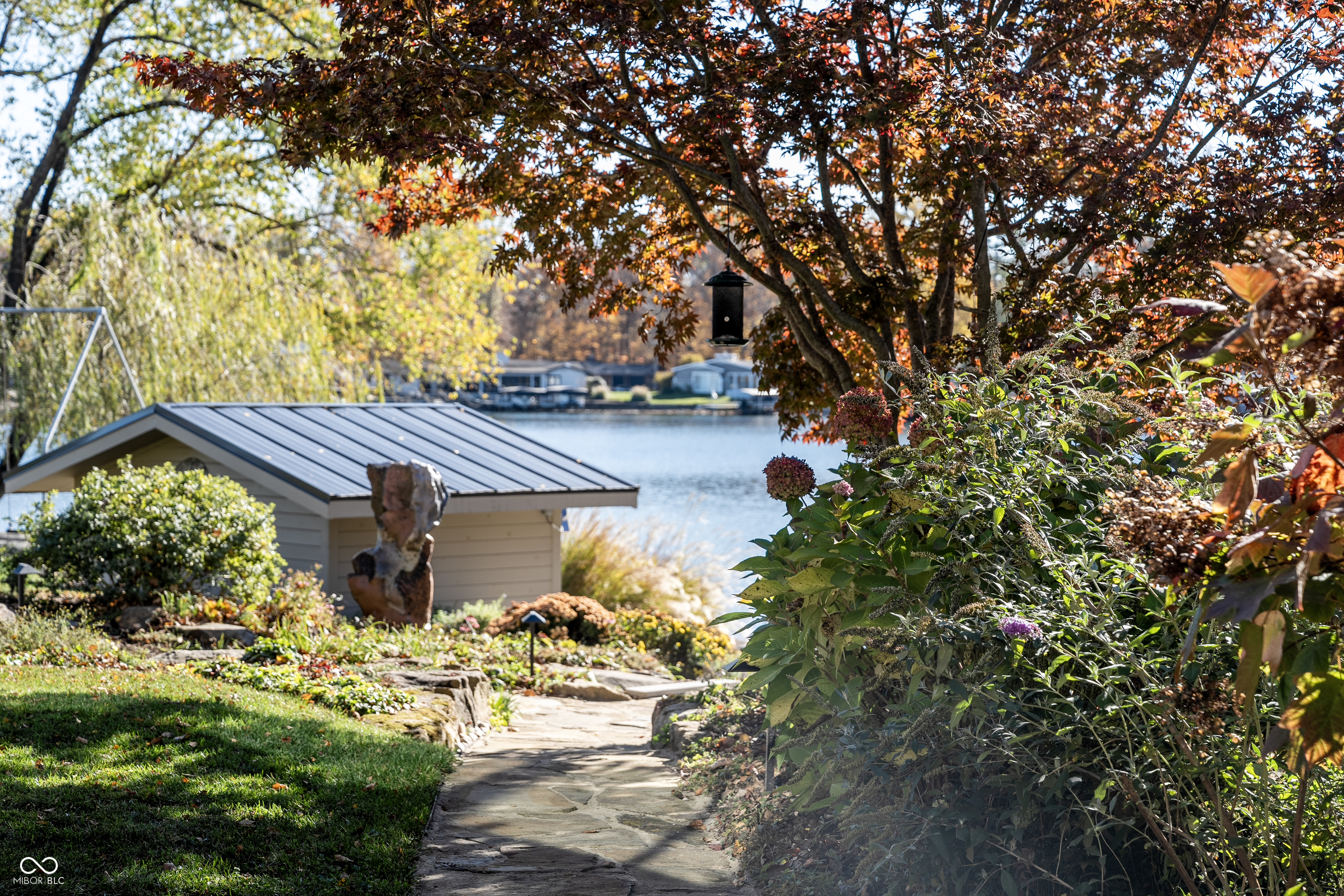 Lakeside backyard with a stone path, flowering shrubs, and an outbuilding beneath autumn foliage.