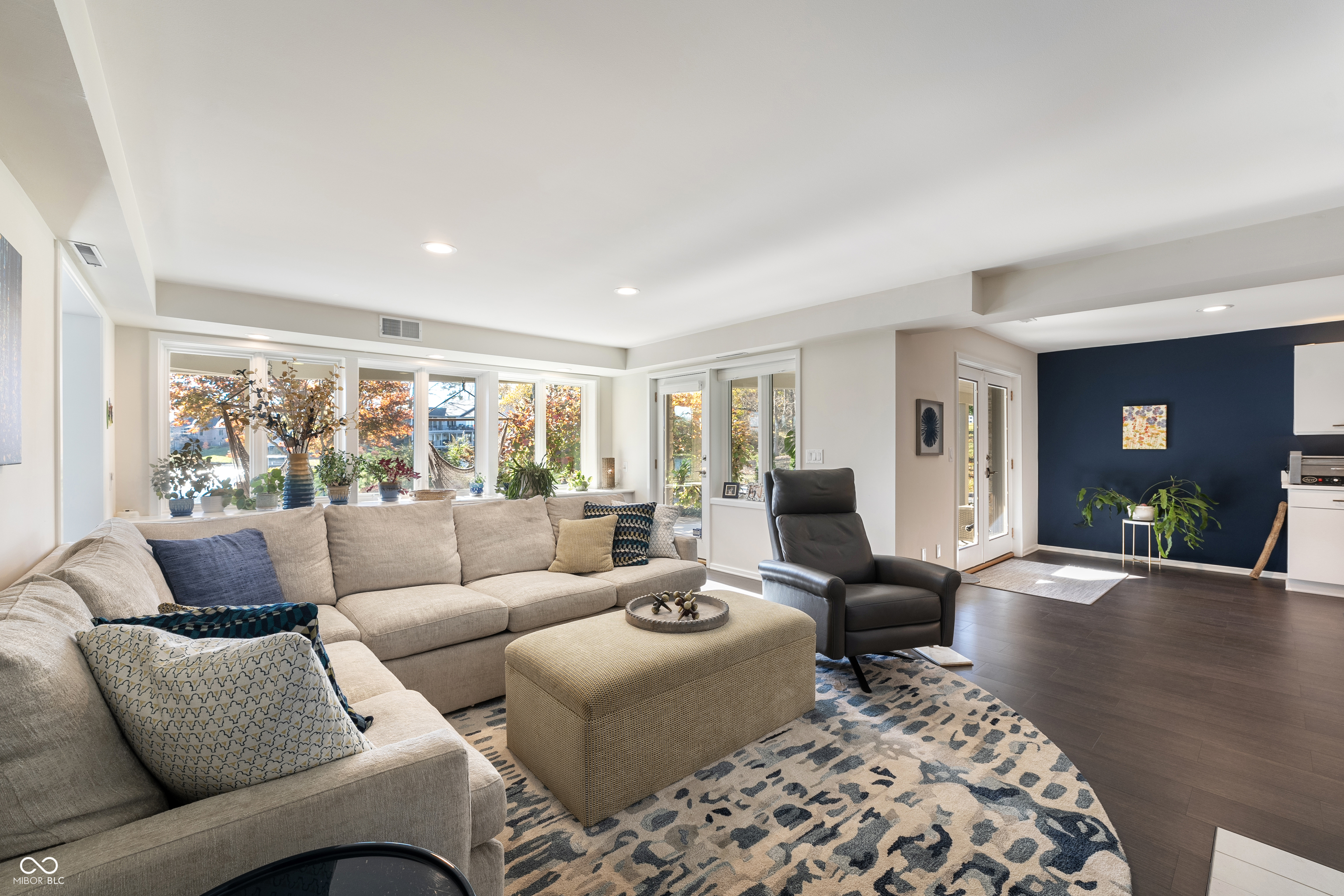 Furnished living room with a beige sectional, patterned rug, and windows overlooking autumn foliage.