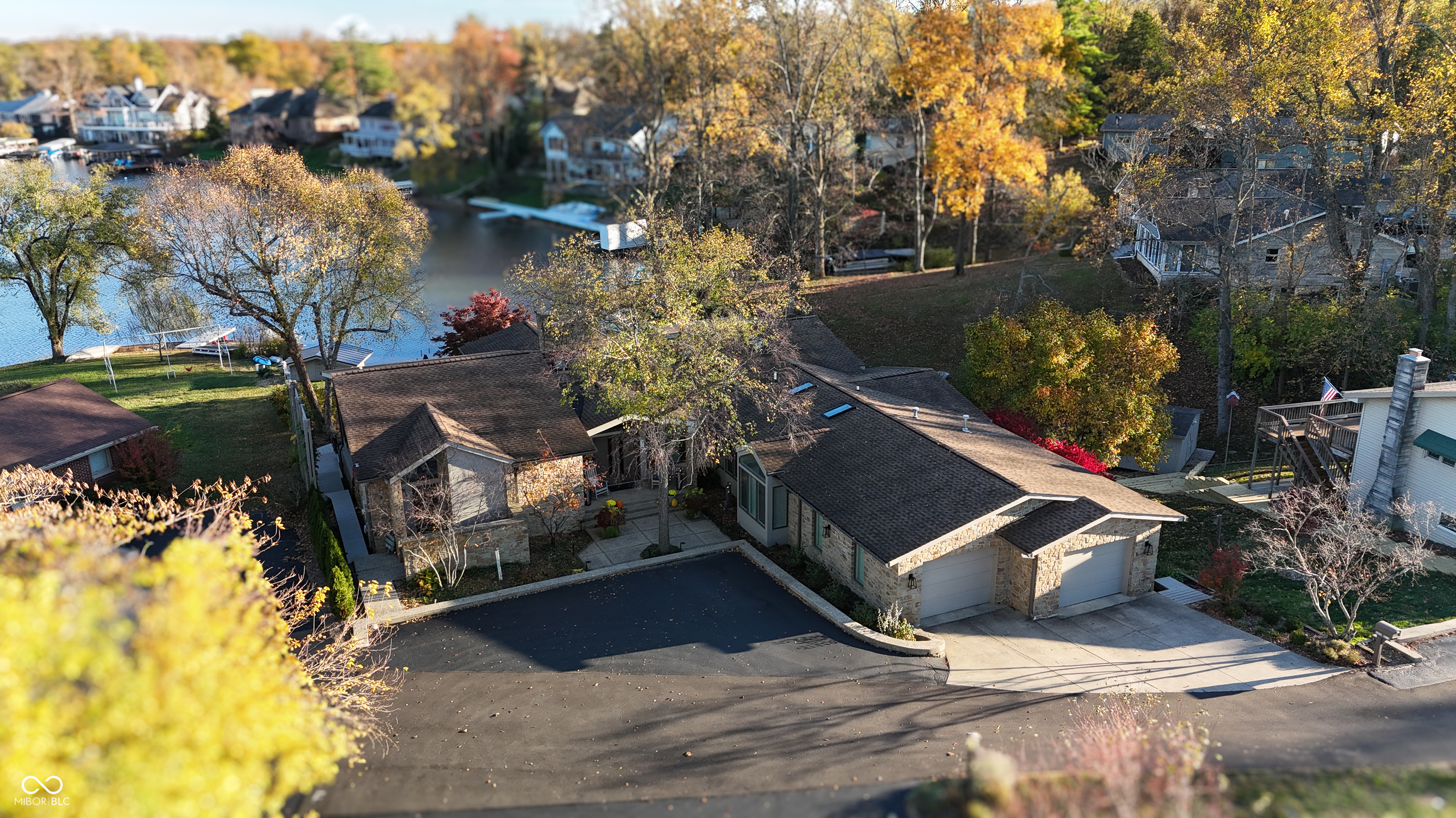 Aerial view of a stone home and curved driveway beside a lake, framed by autumn trees.