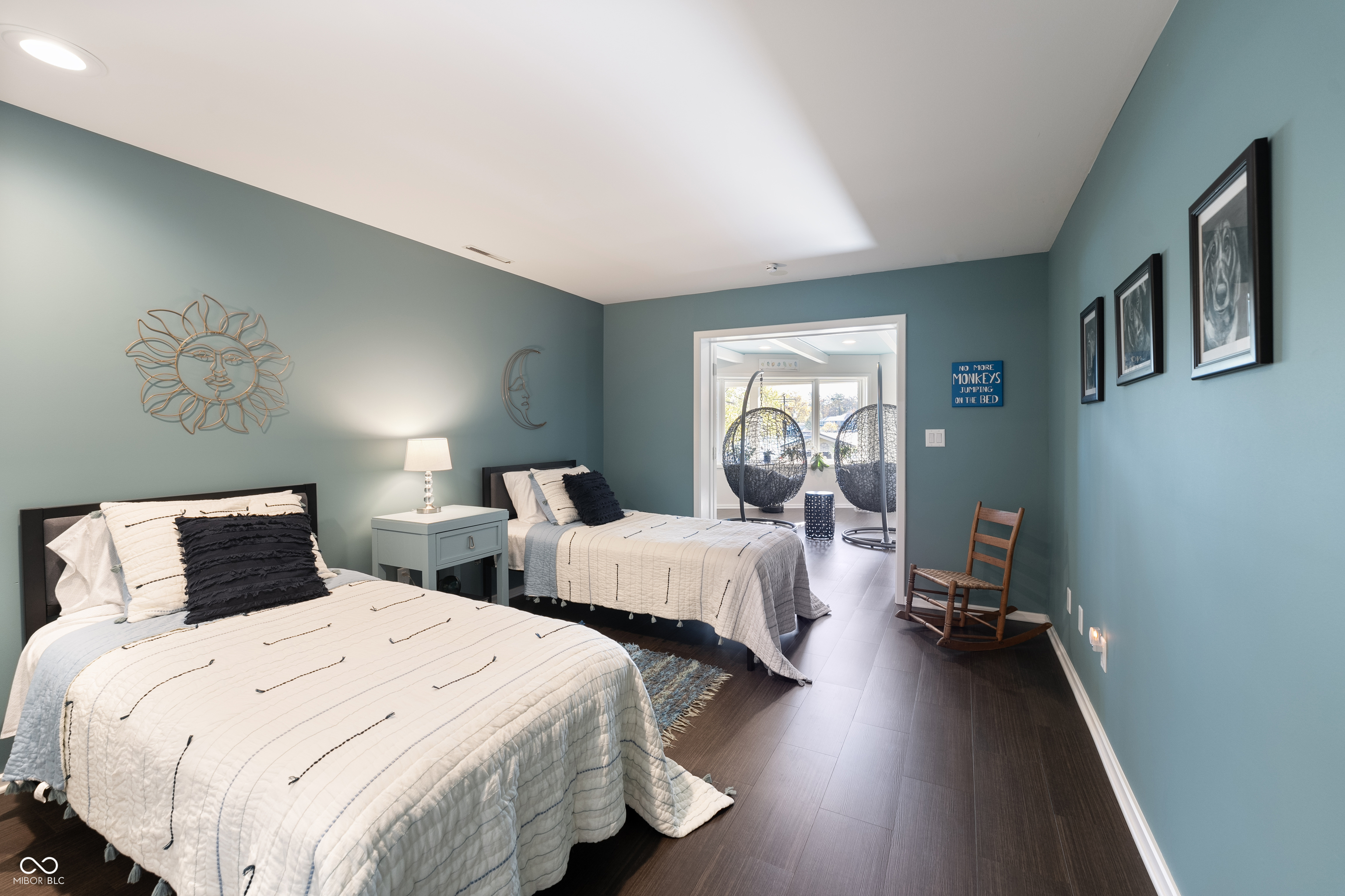 Teal bedroom with two white-covered beds and a doorway to a bright sitting area with hanging chairs.