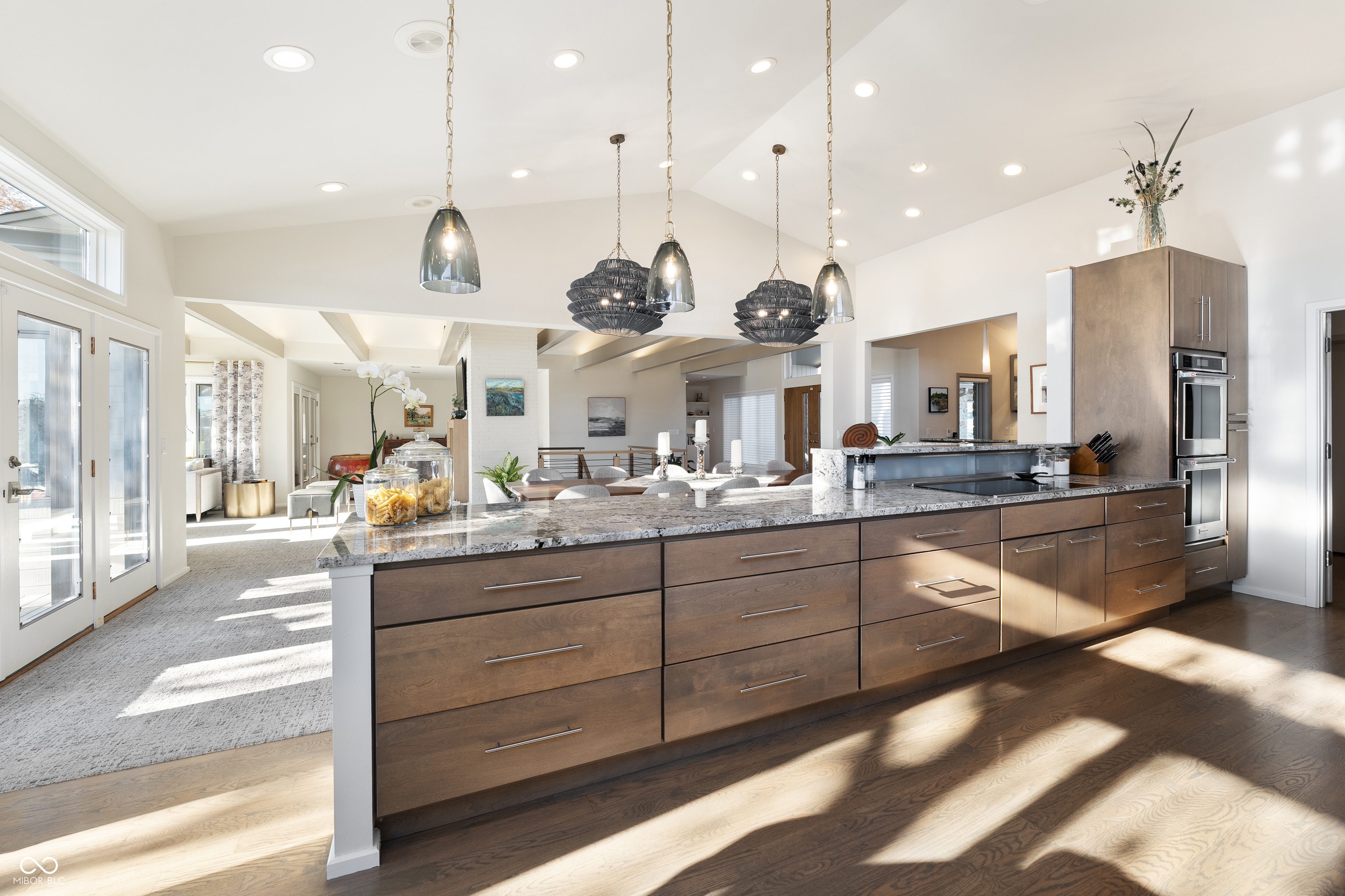 Open-plan kitchen with a large wood-paneled island, stone counters, pendant lights, and double ovens.