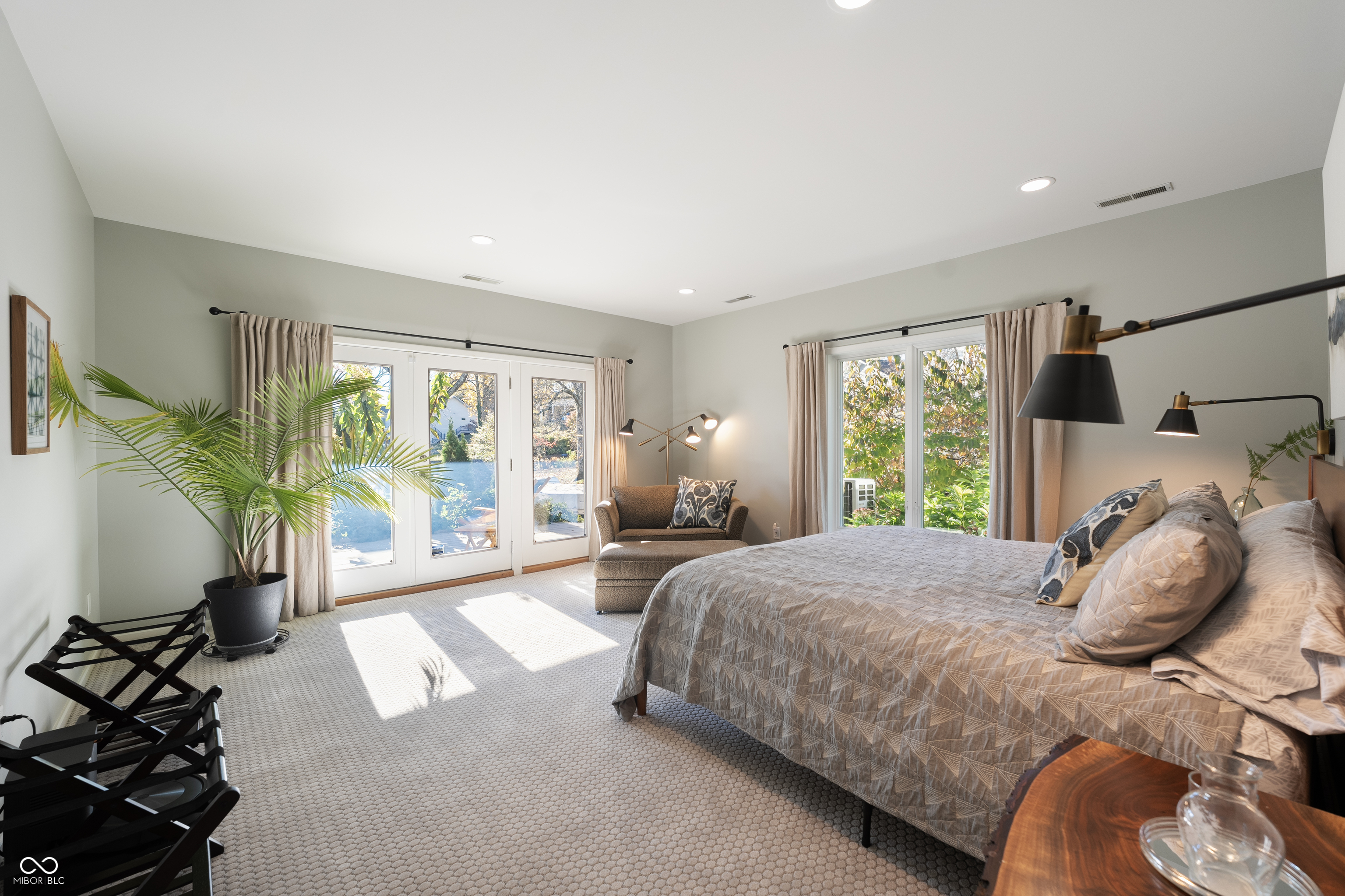 Bright furnished bedroom with a large bed, seating area, and glass doors opening to greenery.