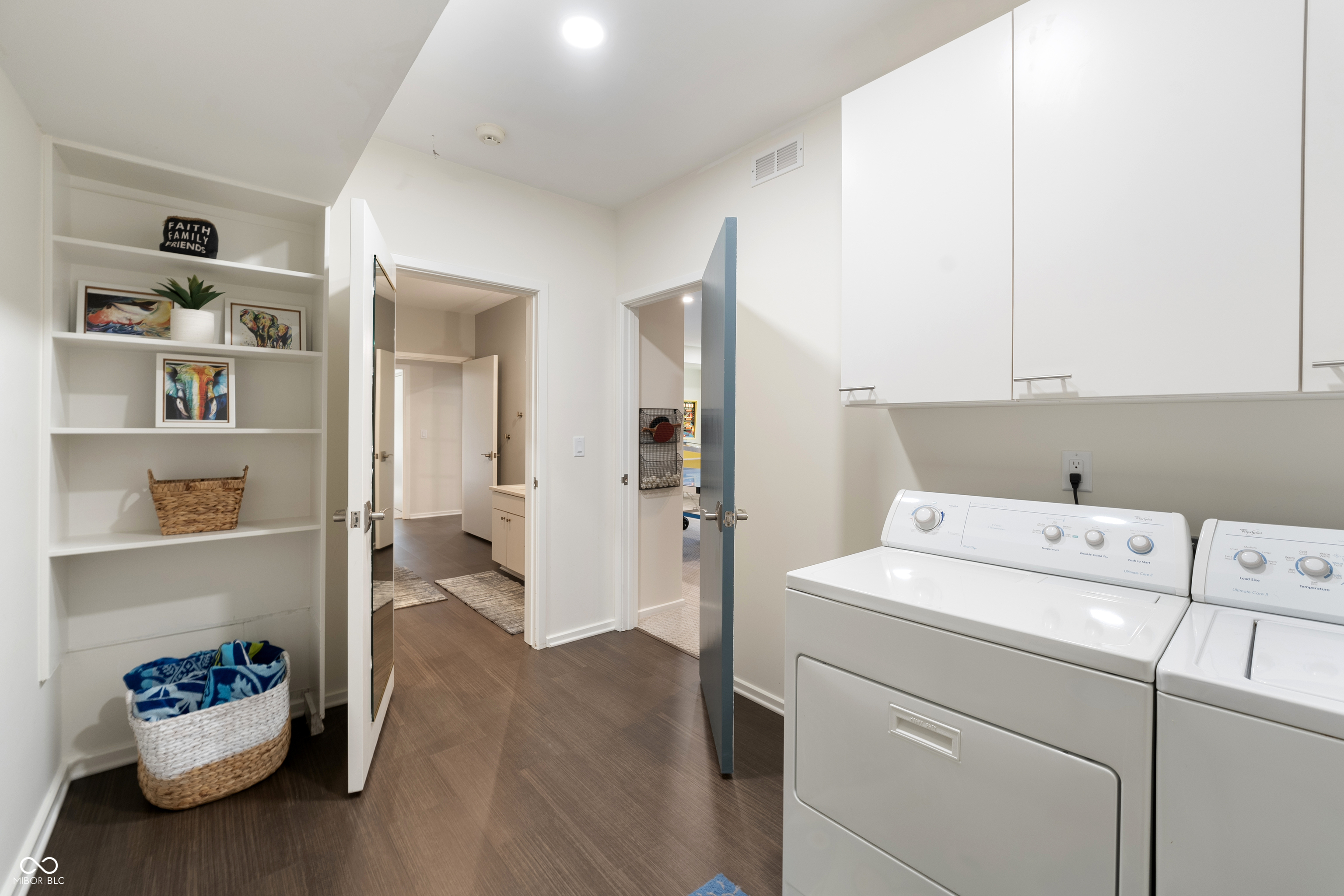Laundry room with white washer and dryer, built-in shelves, and open doors to adjoining rooms.