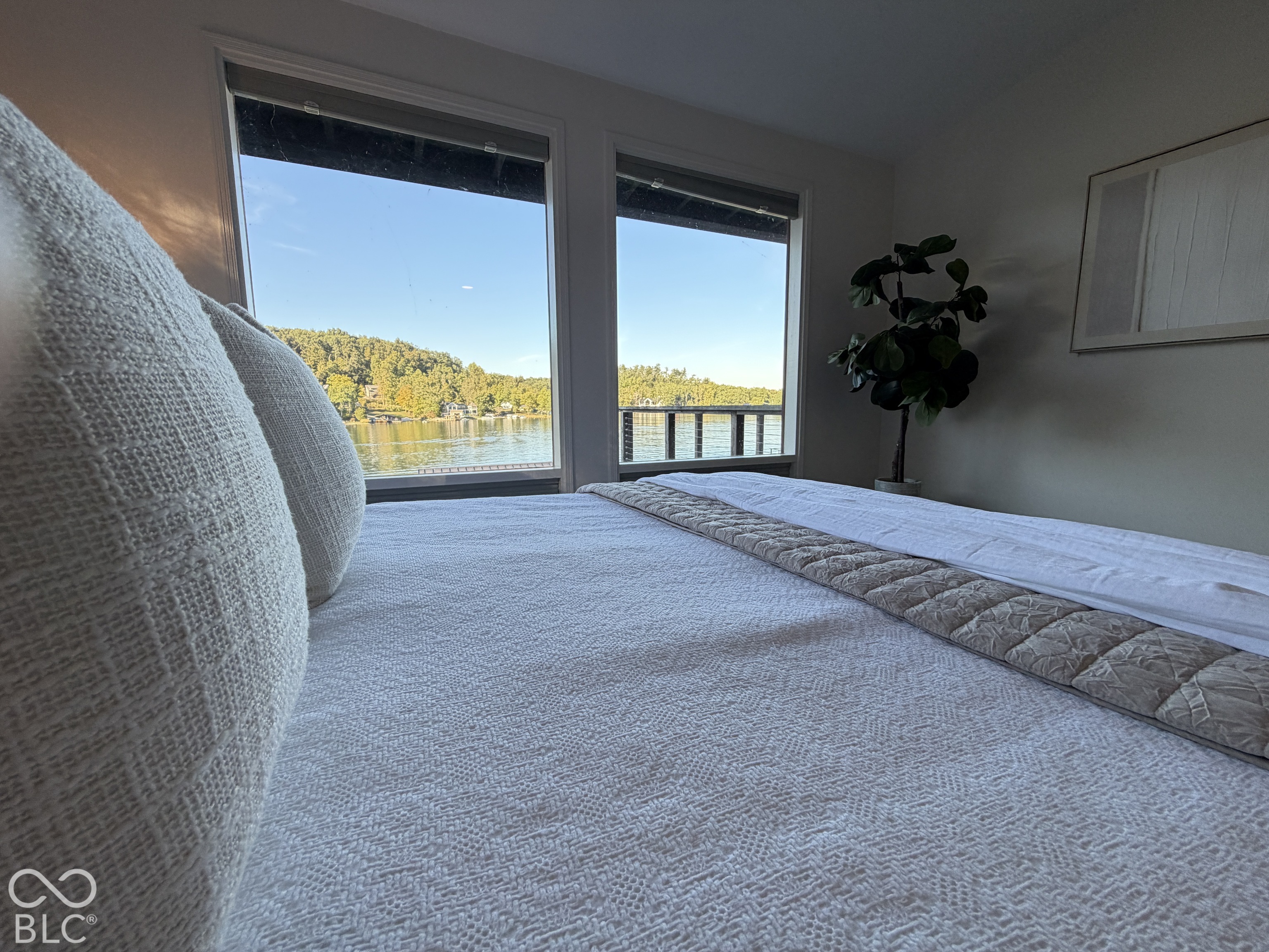 Bedroom with a white bedspread and wide windows overlooking a lake and wooded shoreline.