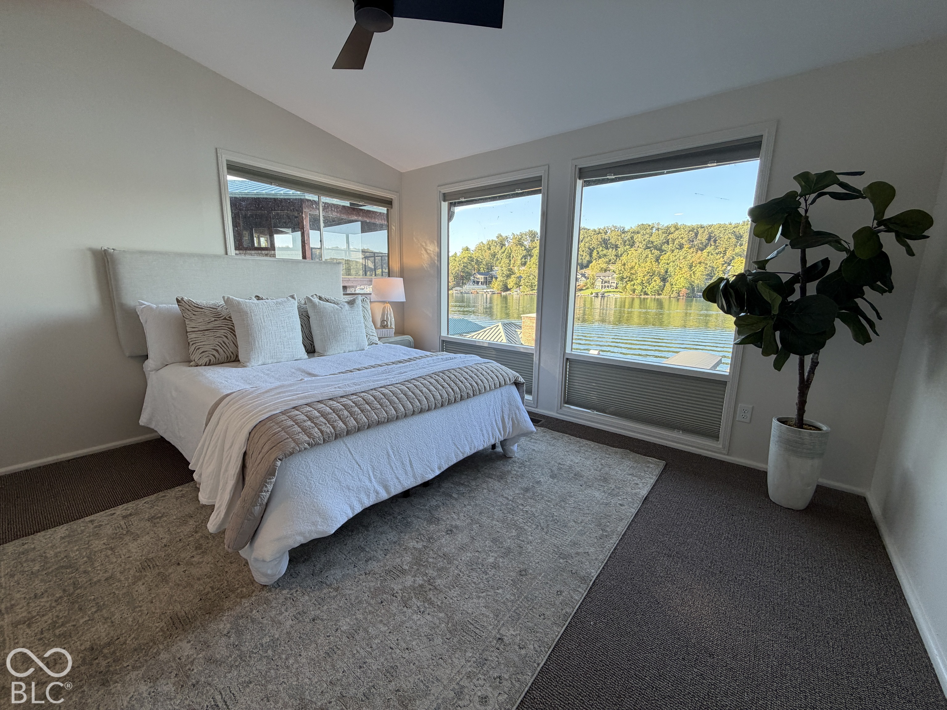 Staged bedroom with a white bed and wide windows overlooking a lake and wooded shoreline.