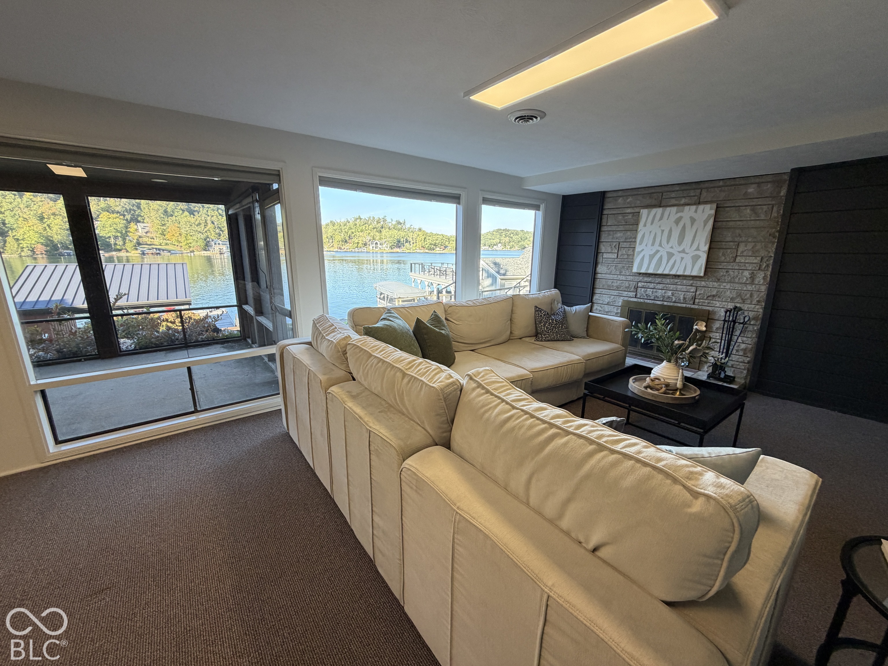 Furnished living room with a beige sectional, stone fireplace, and lake views through large windows.