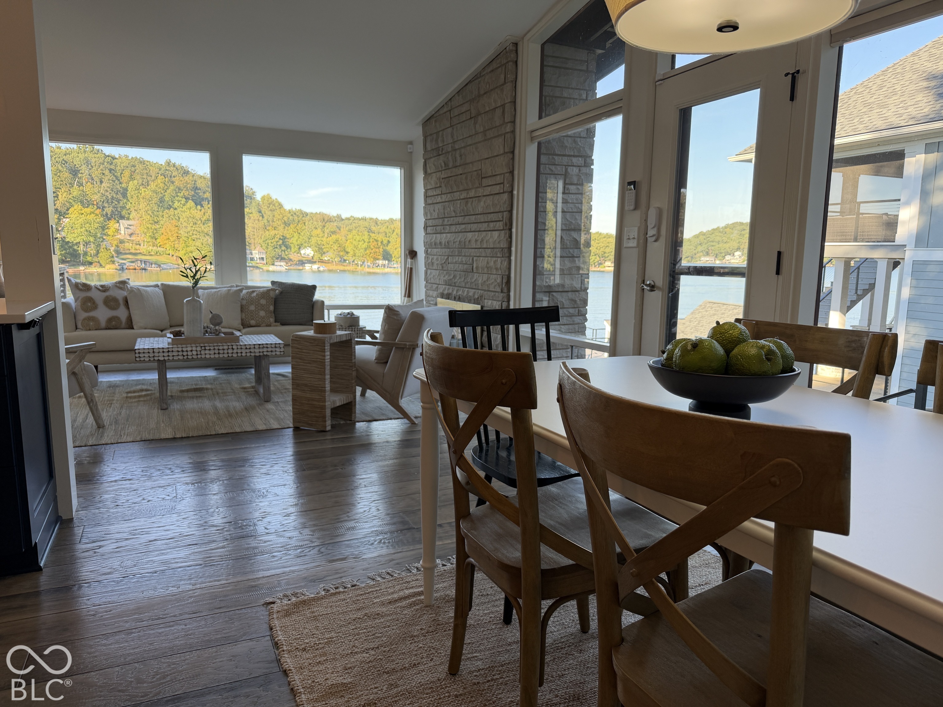 Dining and living area with large lake-facing windows, a white table, wooden chairs, and stone accent wall.