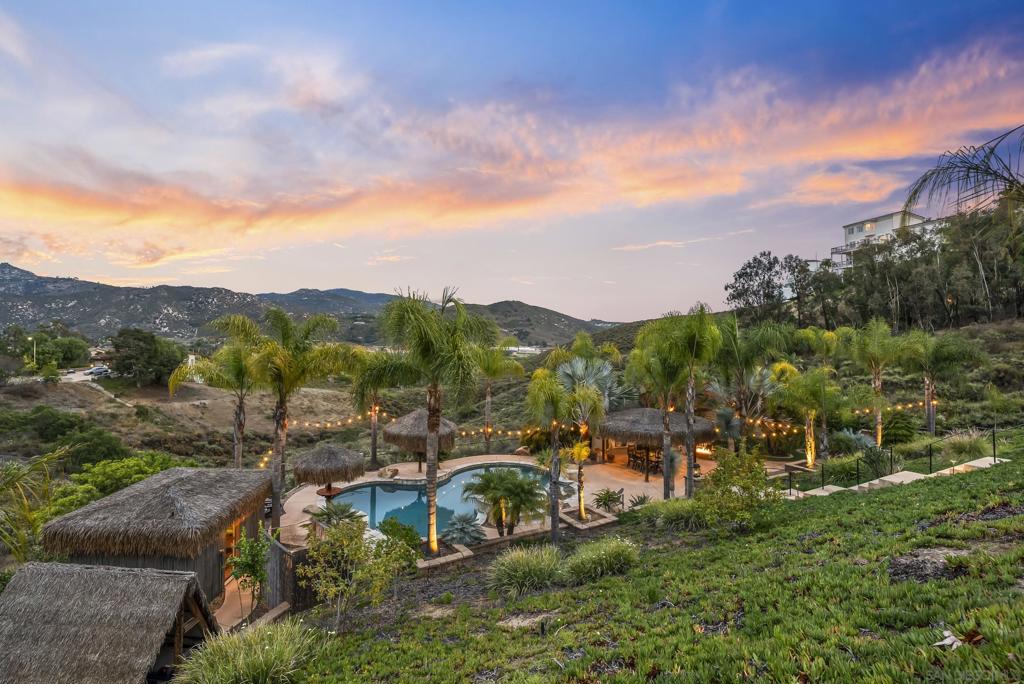 Hillside property with a swimming pool, palm trees, thatched cabanas, and mountain views at sunset.