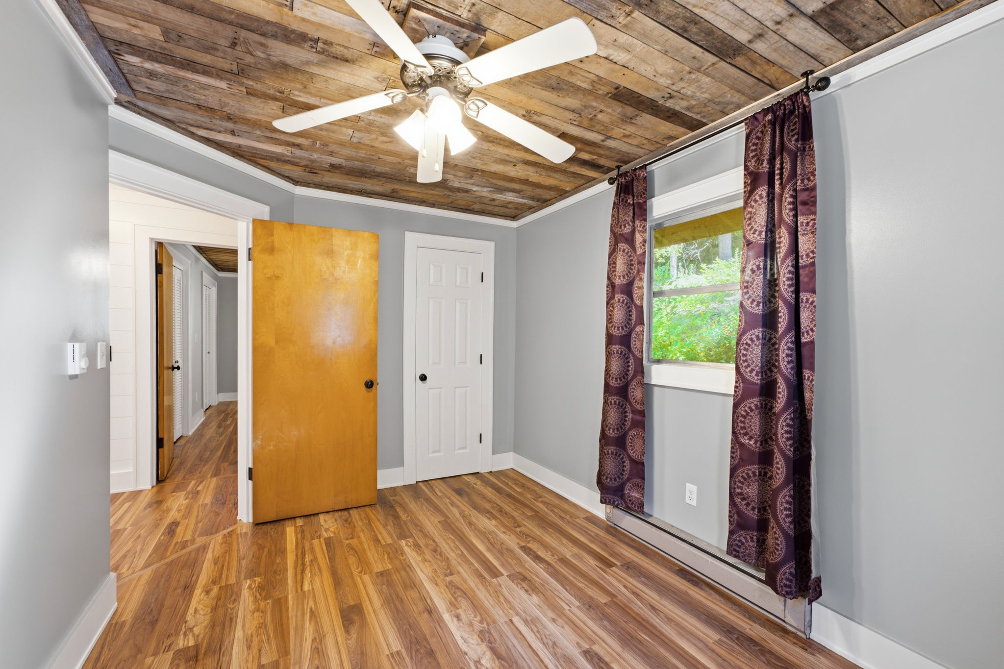 Unfurnished room with a rustic wood ceiling, ceiling fan, patterned curtains, and wood-look floors.