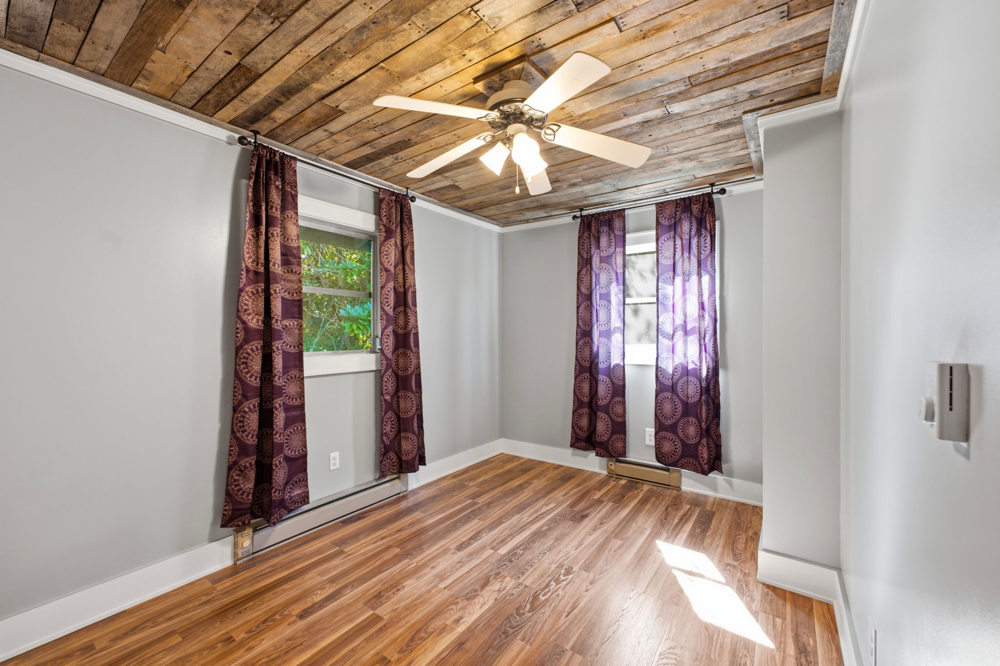 Empty room with a rustic plank ceiling, wood flooring, patterned curtains, and a ceiling fan.