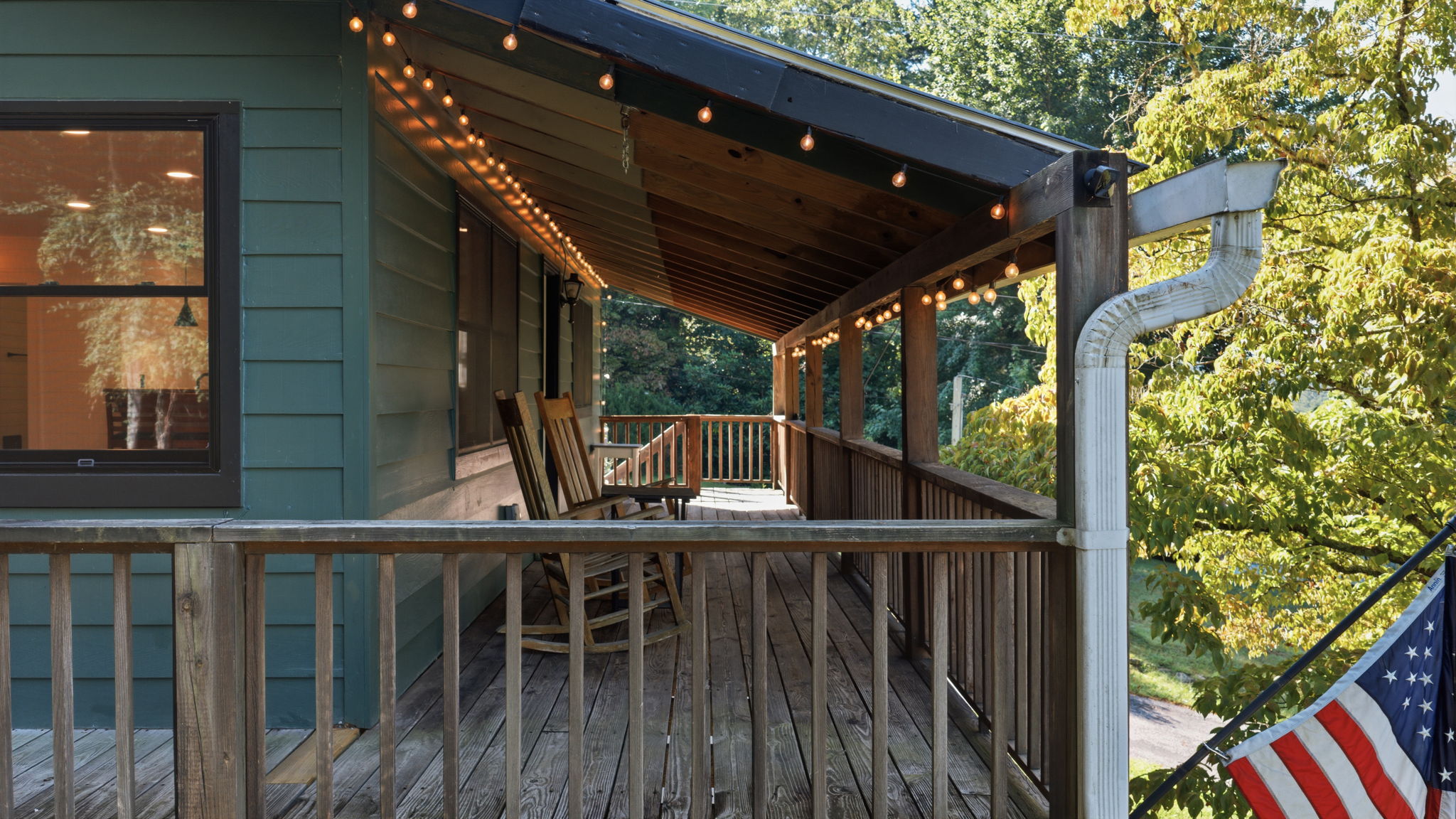 Covered wooden porch with string lights, rocking chairs, and leafy trees beyond the railing.